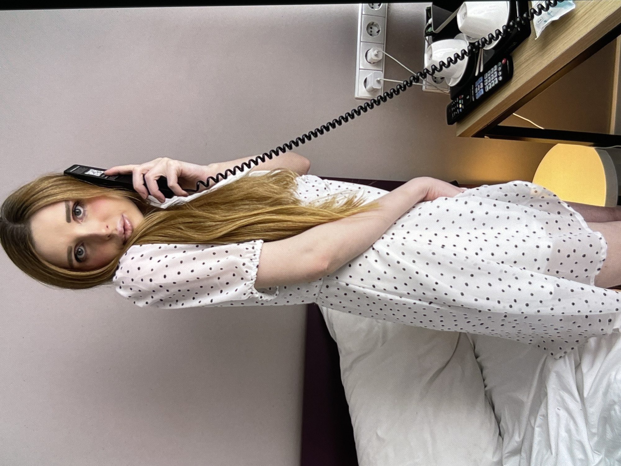 A woman with long, light brown hair is standing in a bedroom, holding a black telephone receiver to her left ear. She is wearing a short-sleeved, white dress with a black polka dot pattern. Her right hand is visible, holding the phone. The background shows a bed with white bedding, a wall, and a small table with a telephone and electrical outlets.