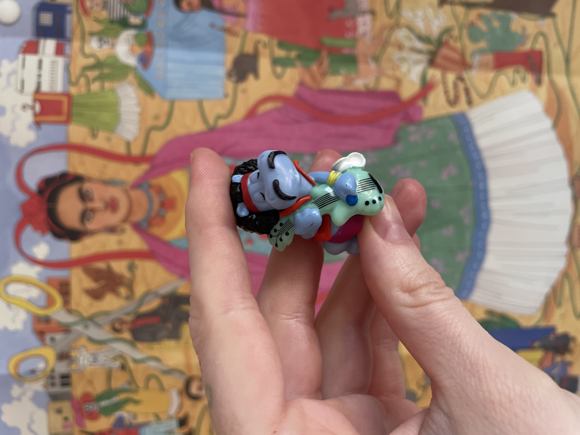 A person holds a miniature figurine in their hand against a background of a colorful jigsaw puzzle, possibly in Vracar Urban Municipality, Serbia. The figurine appears to be a stylized representation of a deity or character, featuring a blue figure lying on its back with what looks like a musical instrument or vehicle-shaped object. The puzzle in the background depicts a collage of images, including what appears to be Frida Kahlo, scissors, and architectural elements. The overall composition suggests a playful or artistic setting.