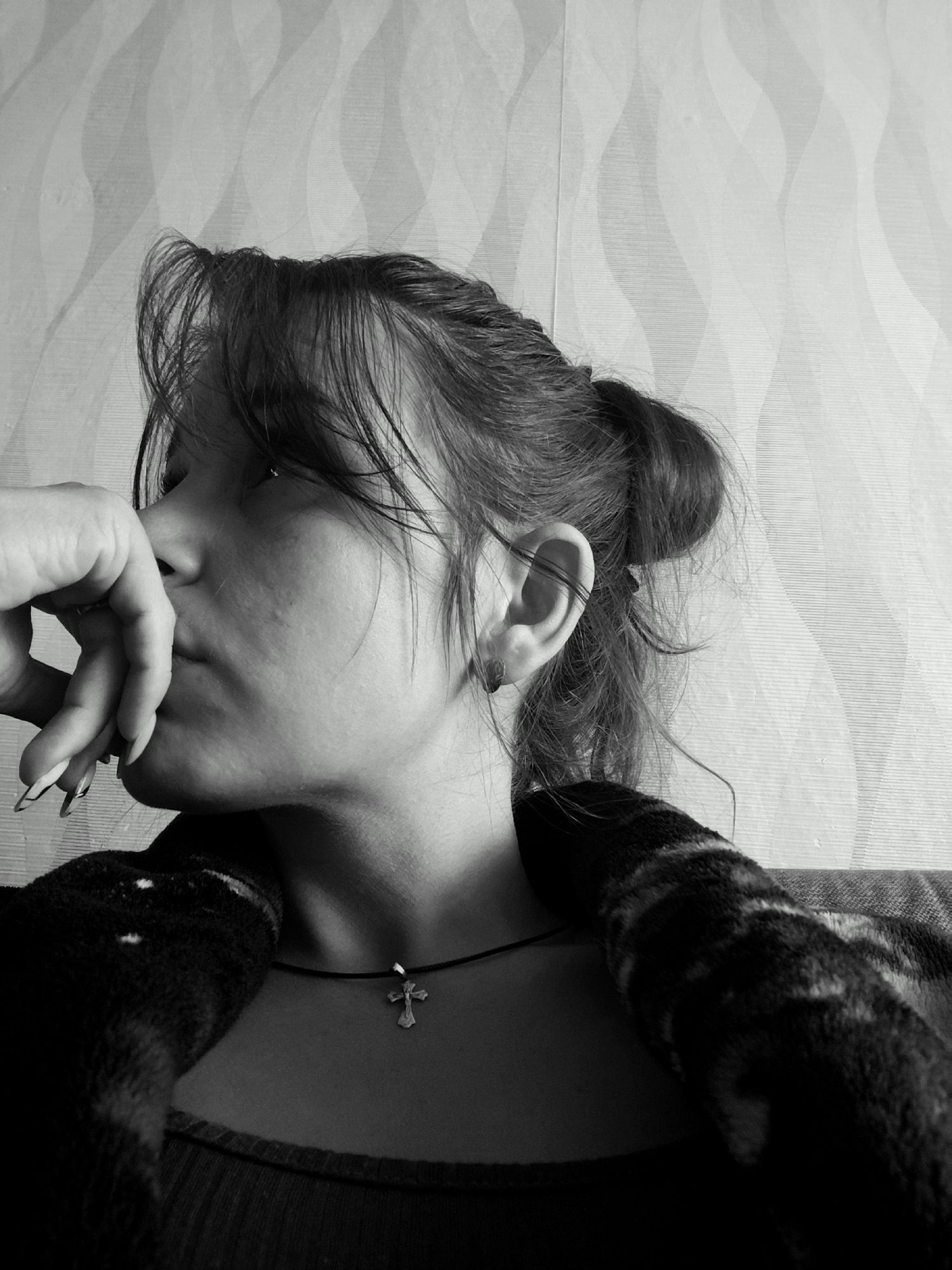 This is a black and white, medium close-up, profile shot of a young woman from the shoulders up. She is resting her chin on her right hand, with her fingers curled in front of her mouth, and her eyes are closed. Her dark hair is pulled back into a bun at the nape of her neck, with some loose strands framing her face. She is wearing a dark, patterned robe or blanket draped over her shoulders, and a dark top underneath. Around her neck, she wears a thin black cord with a small silver cross pendant. The background is a textured wallpaper with a wavy pattern. The lighting appears to be coming from the side, casting shadows on her face. The overall mood is contemplative or somber. There is no visible text in the image. The setting appears to be indoors, likely in a home in Фоки, Russia.