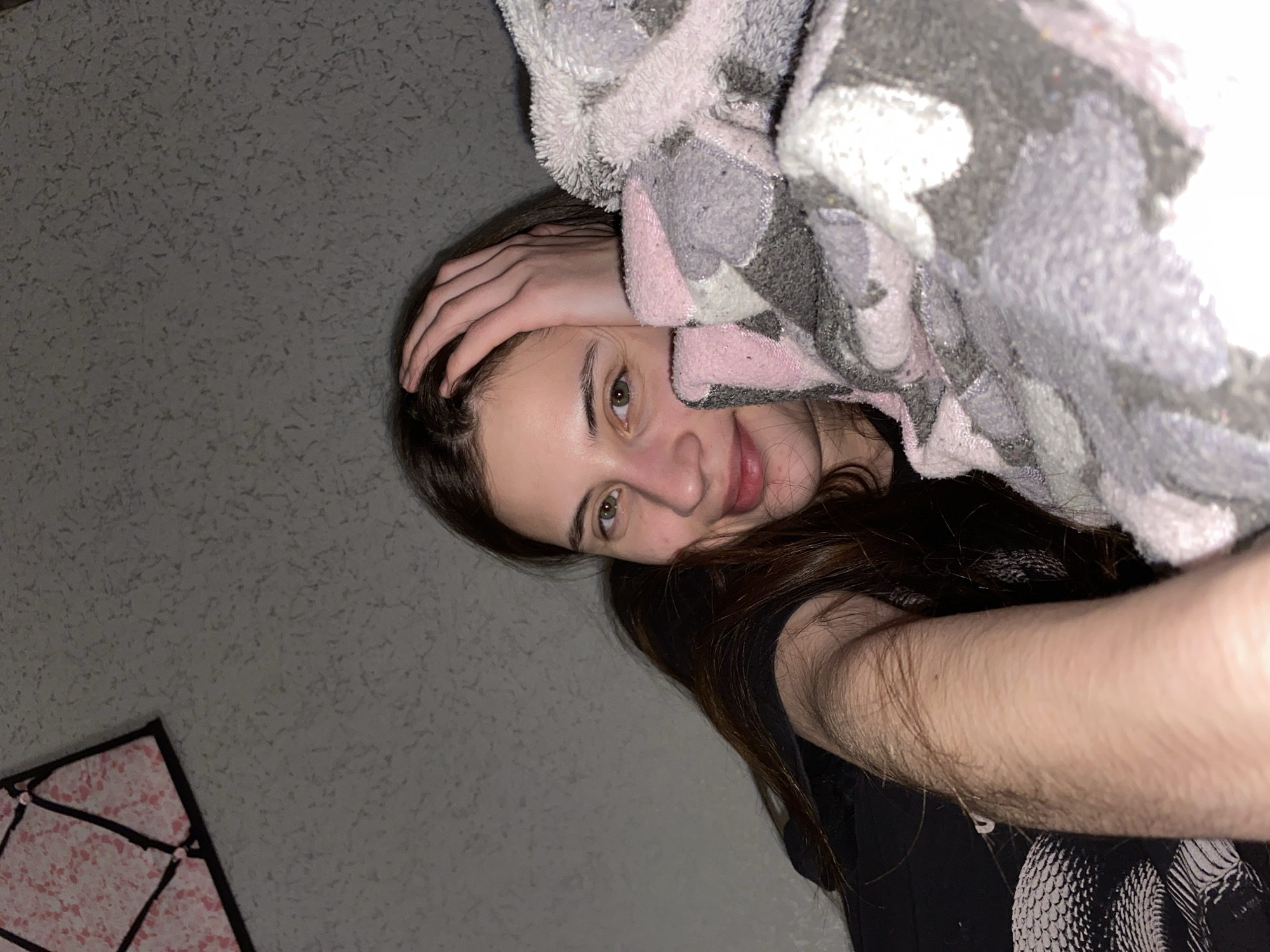 A person is lying down, with their head turned to the right. Their left hand rests on their forehead. A patterned, fuzzy blanket is partially covering the top right of the frame. The person has brown hair and fair skin. They appear to be wearing a black shirt. A patterned object can be seen in the lower left corner of the image, possibly a light fixture or decorative object. The background is a light gray wall. The location of the photo in Votkinsk, Russia is not determinable from the visual information in the picture.