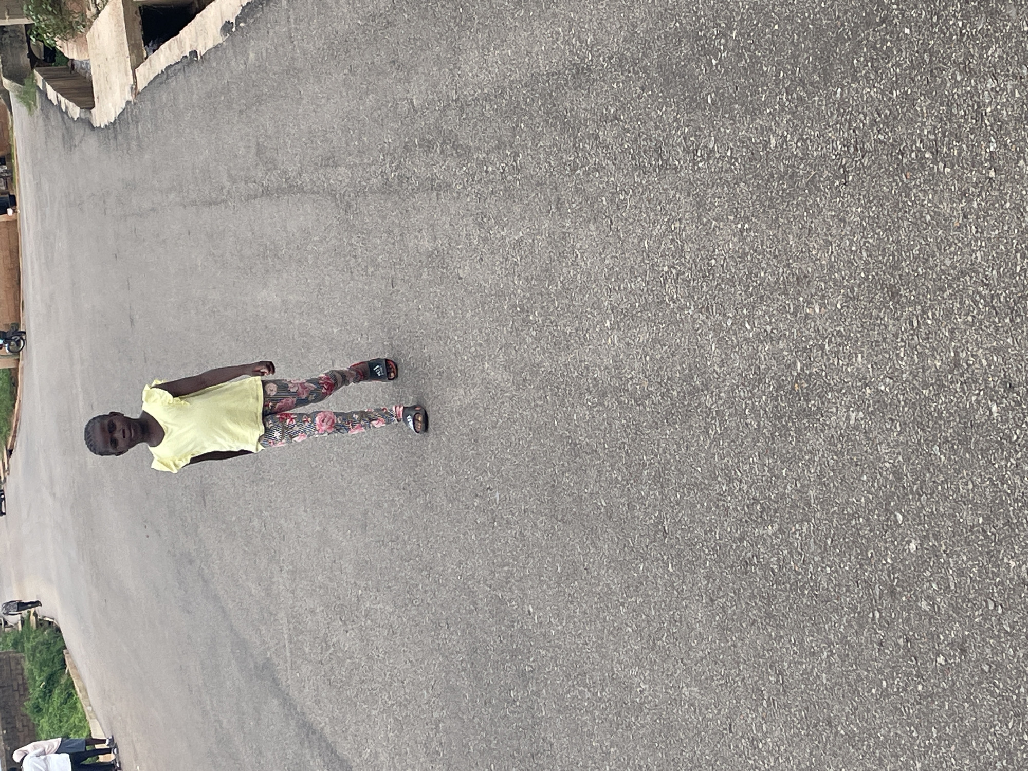 A young child, with dark skin and hair pulled back, stands facing the camera. They are wearing a bright yellow, short-sleeved t-shirt with ruffled details on the shoulders and patterned leggings with a floral design in pink and green. The child is also wearing dark-colored sandals.
The child is standing on a paved, grey asphalt road. To the left of the child, the road slopes downwards and is bordered by a dirt path and some greenery. In the background, there are low-lying buildings with light-colored walls and what appears to be a drainage channel or ditch running alongside the road. Further in the distance, more buildings and vegetation are visible.
The scene appears to be outdoors during the daytime, with natural light casting a slight shadow. The weather seems to be fair. The overall impression is of a child in a residential or semi-rural area. There is no visible text in the image.