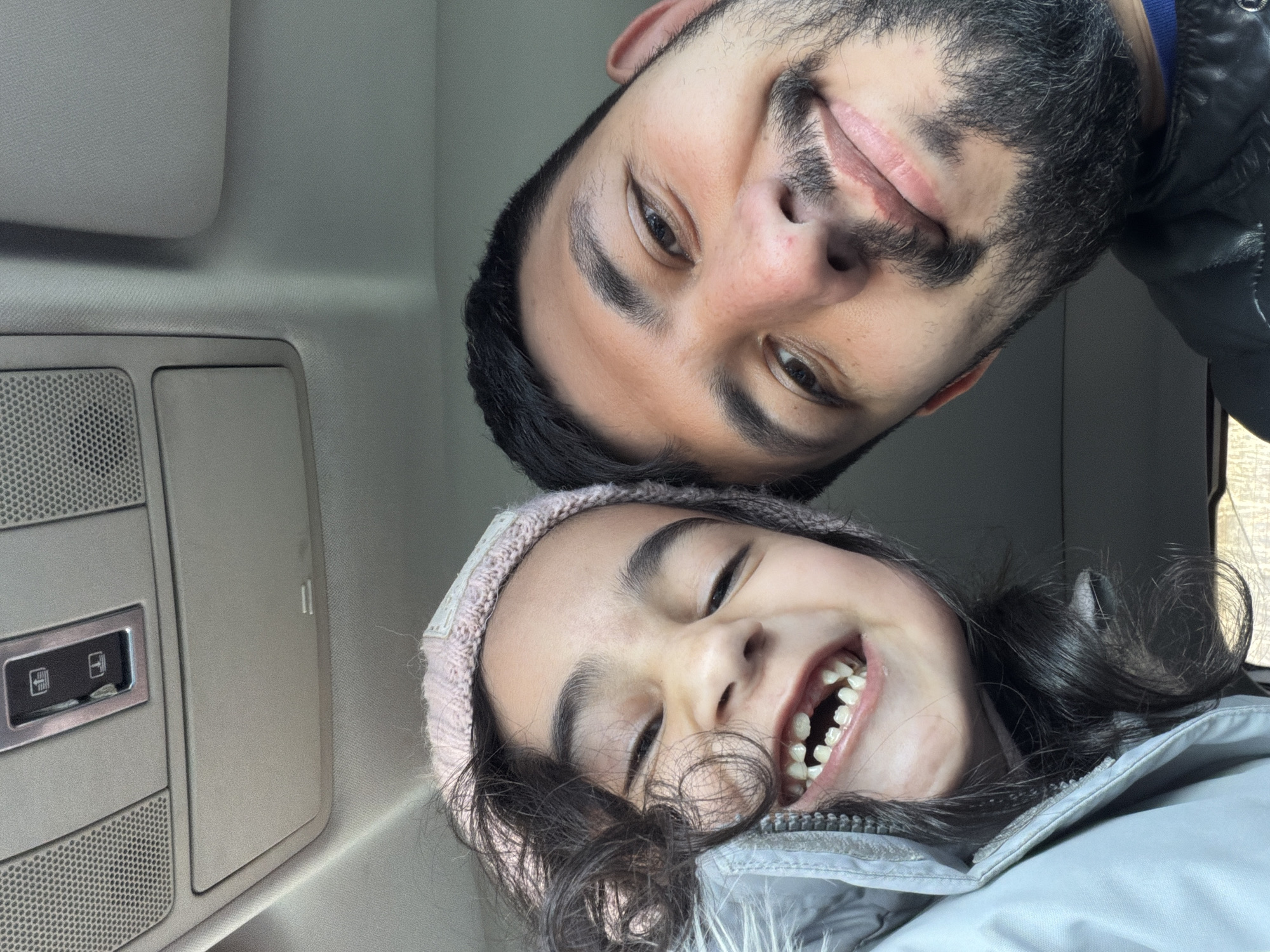 The image captures a selfie of a man and a young girl, likely taken from inside a vehicle, given the overhead console and interior features. The man, with a dark beard and mustache, is positioned above the girl, their faces close together. The girl, wearing a light pink knit hat and a gray jacket, smiles broadly, showing her teeth. The interior of the vehicle includes a gray ceiling and console with visible buttons and vents. The photo appears to be taken at a low angle, tilted to give the viewer an upside-down perspective. The location, Таскино, Russia, cannot be verified from the visual information provided.