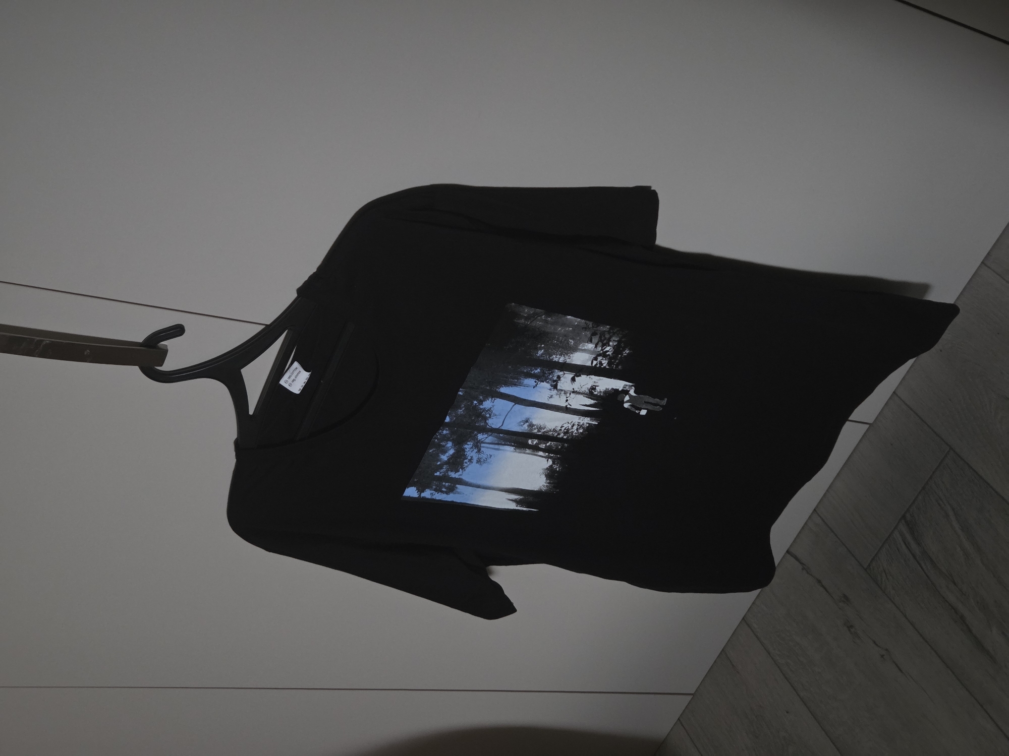 The image displays a black, short-sleeved, crew-neck t-shirt hanging on a black plastic clothes hanger. The t-shirt features a rectangular graphic print on its front, depicting a dense forest scene with dark tree silhouettes against a vibrant blue sky with white, cloud-like formations. A small, stylized figure, appearing to be a person or character, is visible in the lower right portion of the graphic, rendered in light tones and holding a white, rectangular object. The hanger is suspended from a dark, horizontal bar, which is mounted on a light grey vertical panel, likely a wall or part of a closet unit. A white textile tag, partially visible on the t-shirt's interior collar, indicates "100% cotton" and "100% Baumwolle." The immediate background consists of plain, light grey walls or closet doors, with a section of light-toned wooden flooring, arranged in a herringbone pattern, visible in the lower left of the frame. The location is an indoor setting, likely a room within an unknown building in the city of Unknown, Unknown. No discernible actions or interactions are taking place.