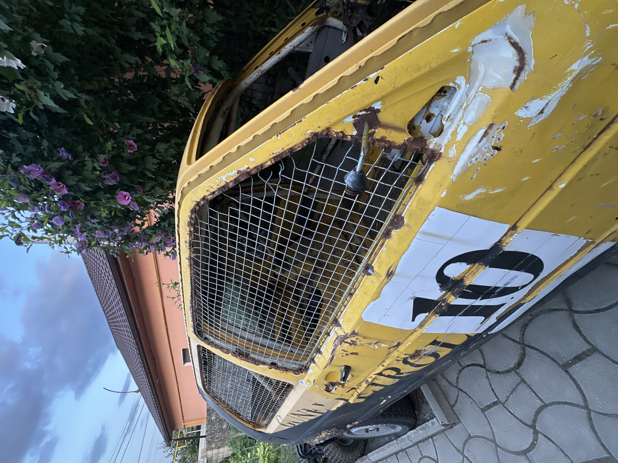 An abandoned, rusty yellow car with black accents and the number "10" in large white lettering on its side is parked on a tiled surface next to a building with an orange facade and a brown roof. The car's windows are covered with a wire mesh, and the paint is peeling and shows signs of significant wear. Green foliage with purple flowers partially obscures the car and the building. The scene appears to be an outdoor setting in a residential area, potentially Comrat, Moldova, given the architectural style and vegetation. The sky above is overcast with gray clouds. There are no people visible in the image, and the car shows no signs of recent activity or use.