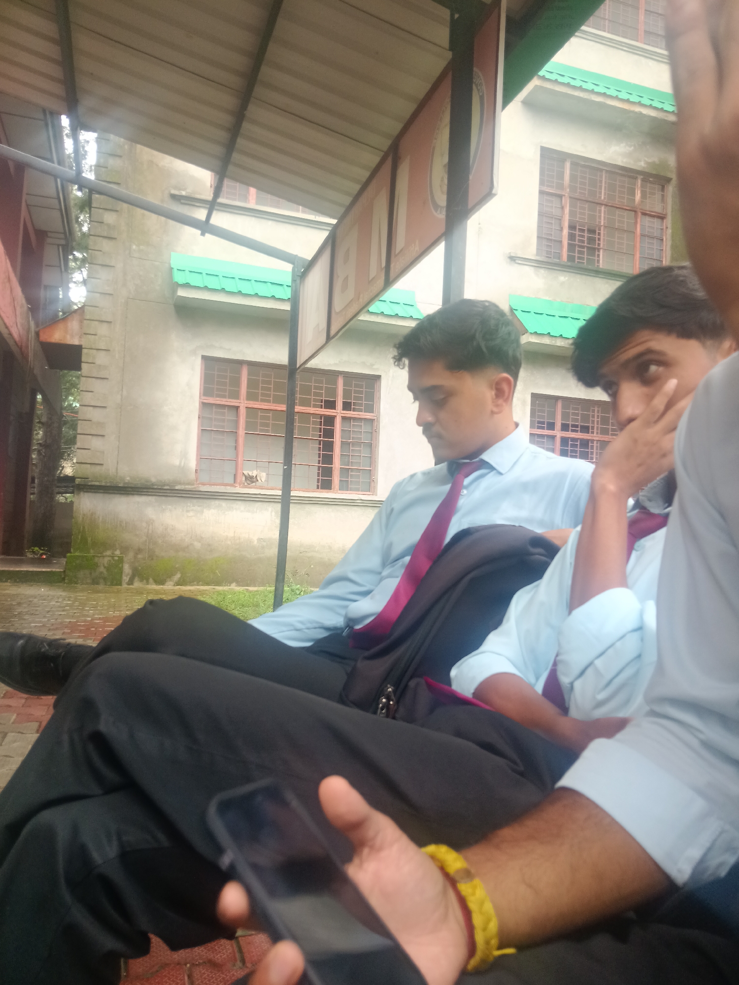 A medium shot captures three young men in light blue collared shirts and dark ties, seated outdoors under a corrugated metal awning. The man in the foreground, on the right, looks away from the camera, his hand near his face. Another man sits to his left, looking downwards. The third man is mostly obscured by the other two and an awning. All three appear to be wearing black trousers. The background shows a beige building with green trim and multiple windows. The ground is paved with red bricks and some grass. This scene is located in Dharamshala, India, a city known for its Tibetan Buddhist culture and scenic mountain views.