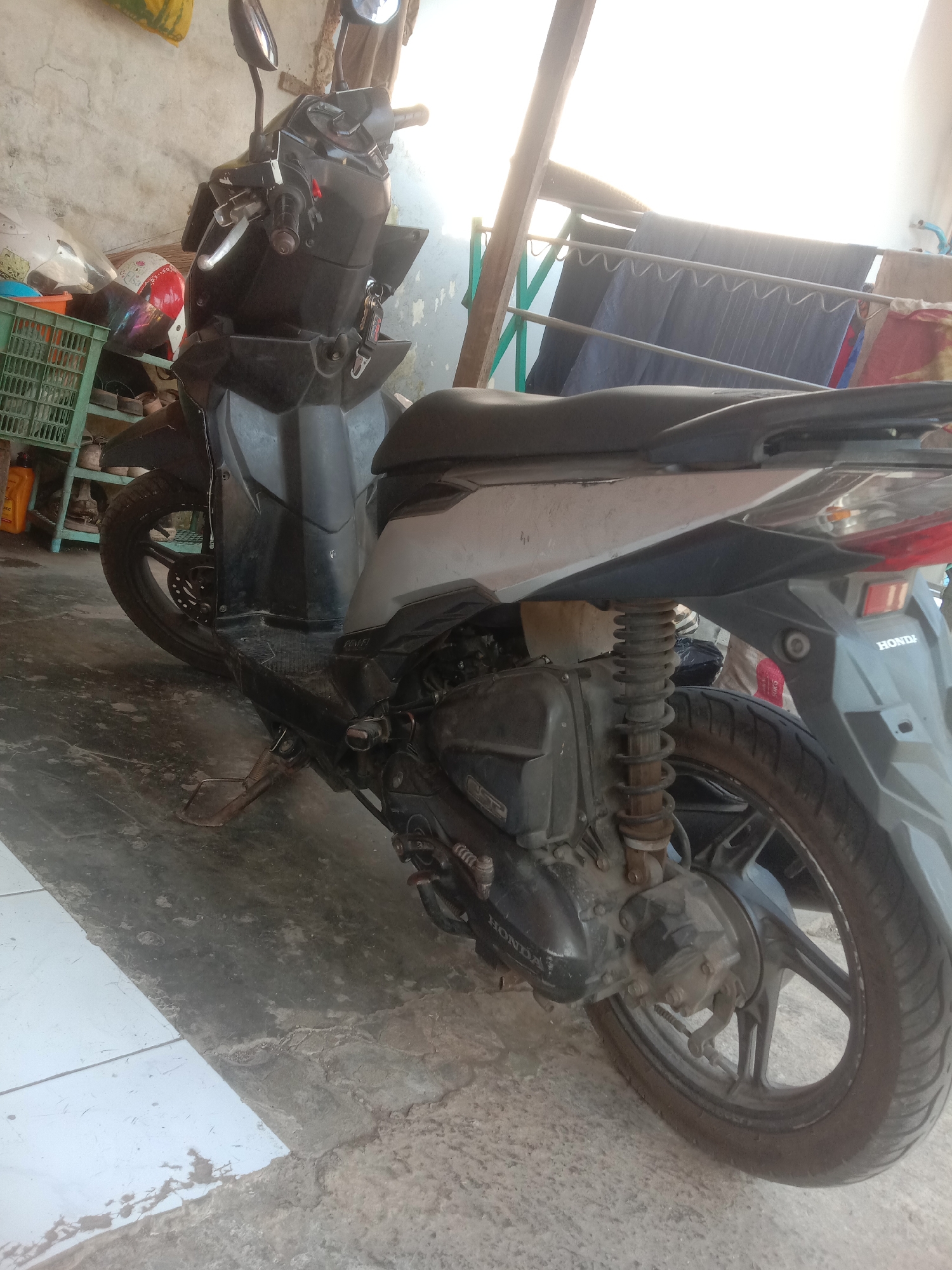 An outdoor scene in Cianjur, Indonesia, features a black scooter parked on a concrete floor. The scooter's rear is angled towards the right of the frame. In the background, a makeshift clothes rack holds several dark-colored garments. To the left of the scooter, a green plastic crate is visible, containing what appears to be a helmet and other miscellaneous items. The environment suggests a residential or workshop area, with rough walls and a somewhat cluttered appearance. Sunlight streams in from the upper right, casting a bright glare.