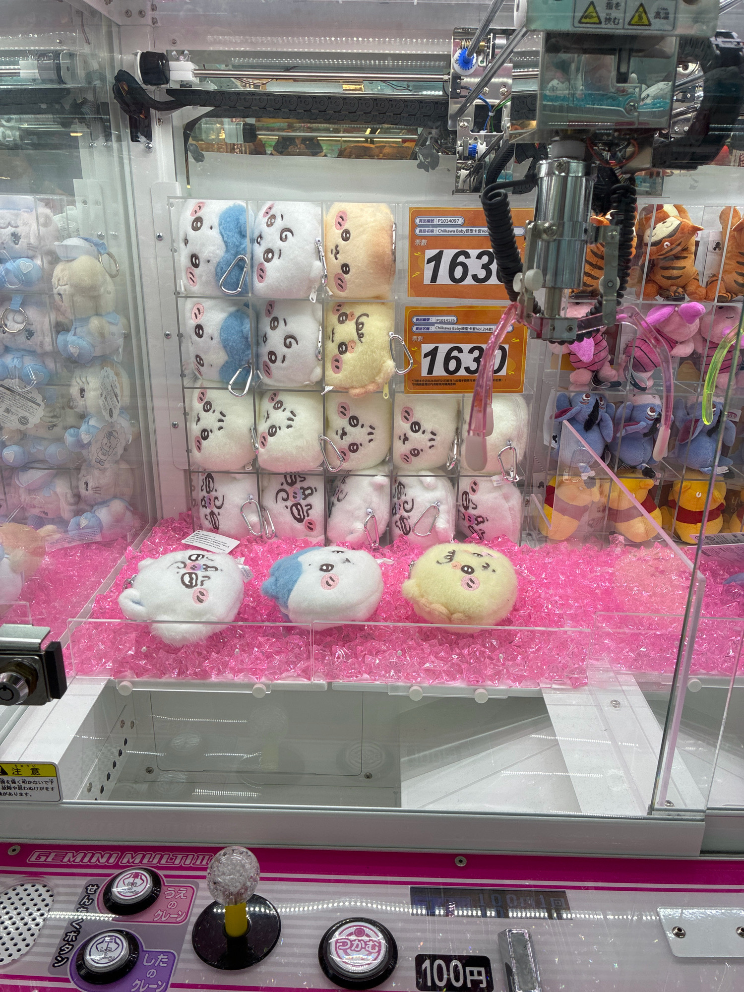 The image shows the interior of a claw machine arcade game in Hong Kong, China. The machine is filled with various plush toys, including small, round, character-faced stuffed animals in pastel colors such as white, blue, and yellow, as well as larger stuffed animals resembling tigers and characters from Winnie the Pooh. The plush toys are arranged on multiple shelves and in the main play area, which is covered in pink, crystalline filler material. The claw mechanism, a metallic gripper with a pink plastic covering, is visible at the top of the machine, poised above the toys. Pricing information and product descriptions, written in Japanese and Chinese, are displayed on orange and yellow signage. The exterior of the machine features control buttons and a coin slot, indicating it is a coin-operated game. The overall environment appears to be an indoor entertainment facility, typical of an arcade.