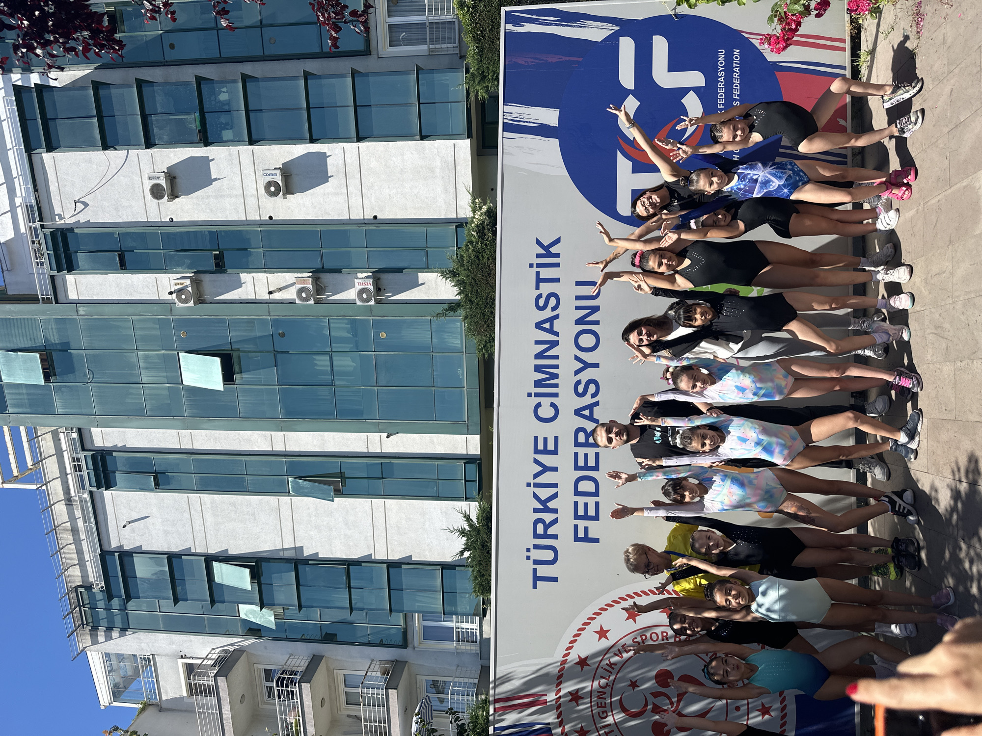 In Sancaktepe, Turkey, in front of a modern building with multiple stories and blue-tinted windows, a group of young gymnasts stands with their hands raised in a celebratory pose. The building's facade features several air conditioning units and balconies. A large banner behind the gymnasts displays the text "TÜRKİYE CİMNASTİK FEDERASYONU," which translates to "Turkish Gymnastics Federation." The banner also includes the logo of the federation, a stylized "TCF" within a blue circular emblem. The gymnasts are wearing various leotards, some plain and others with colorful patterns. Two adults, likely coaches, are positioned behind the children, also participating in the pose. The ground is paved with light-colored tiles, and to the right of the image, a pink flowering bush is visible. The sky is clear and blue, indicating a sunny day.
