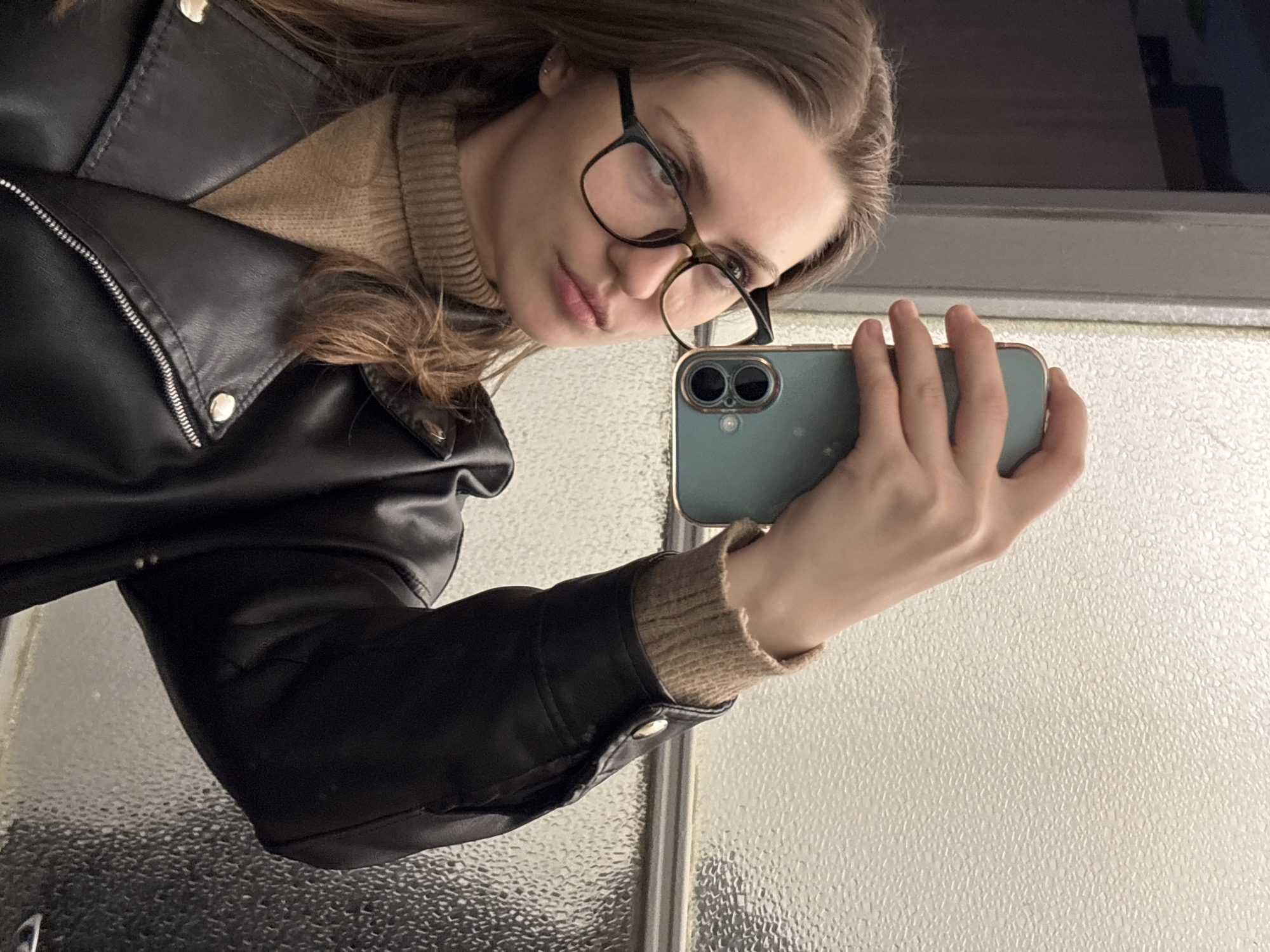 A young woman is taking a selfie in a room with a textured glass wall or window. She is wearing black-rimmed glasses, a tan turtleneck sweater, and a black leather jacket. The phone she is holding is blue and has two camera lenses. The location is unknown, and there is no evidence to indicate that the picture was taken in Saint Petersburg, Russia.