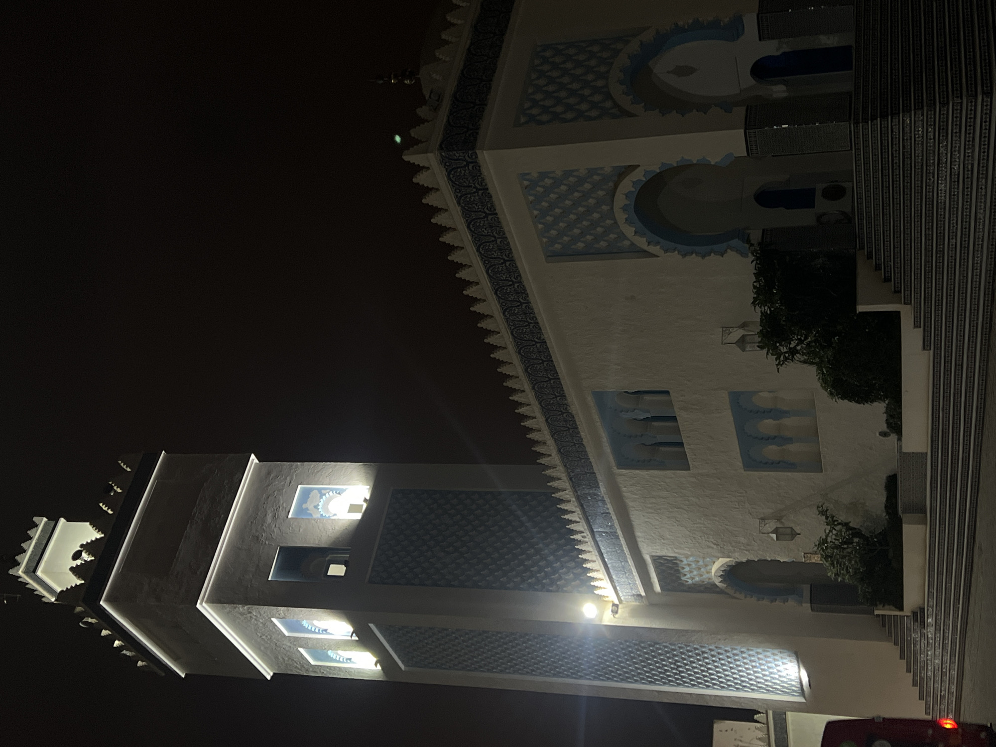 The image displays an ornate white building, likely a mosque or a structure influenced by Moroccan architecture, illuminated at night in M'Diq, Morocco. The building's facade is adorned with intricate blue and white geometric patterns, including diamond-like textures and decorative friezes along the roofline and window borders.
On the left side, a tall tower or minaret rises, featuring crenellated tops and illuminated sections that highlight its architectural details. The main body of the building also has multiple arched window frames and decorative panels with similar blue and white motifs. Bright artificial lights are visible within some windows and on the exterior, casting a warm glow that contrasts with the dark, featureless night sky.
Towards the right, a section of stairs with patterned risers ascends, flanked by dark green foliage. A small part of a vehicle, identifiable by its red taillight, is visible in the bottom right corner. The overall scene emphasizes the building's distinctive architecture and its illumination against the darkness, suggesting a calm evening or night setting. The image is oriented sideways (rotated 90 degrees counter-clockwise).