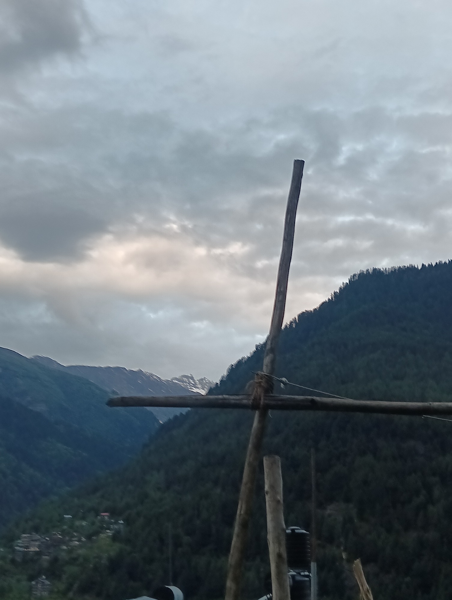 The image depicts a mountainous landscape with a cloudy sky. In the foreground, there are wooden poles and some rudimentary scaffolding structure. The background features a steep, tree-covered mountainside, and further in the distance, more mountains with snow-capped peaks are visible. A valley with a small settlement and scattered houses can be seen nestled between the mountains. The lighting suggests it might be dusk or a heavily overcast day. The overall impression is one of a remote, natural setting, potentially in a rural area like Vashisht, India, known for its mountain views and Himalayan scenery.