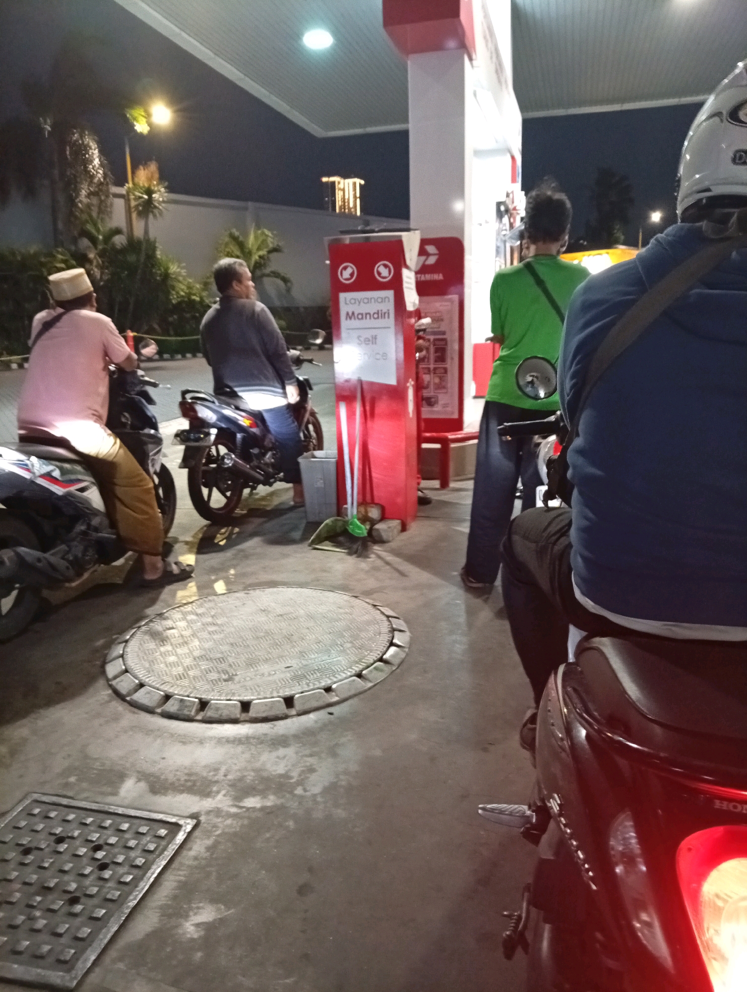 The image depicts a scene at a Pertamina gas station in Surabaya, Indonesia, during the evening. Several individuals on motorcycles are present, either waiting or having just filled up. In the foreground, a person wearing a blue jacket sits on a red motorcycle, facing away from the camera. To the left, another individual in a pink shirt and a white cap is seated on a motorcycle. Behind them, a person in a gray jacket is also on a motorcycle, looking towards the gas pumps. Further back, a person in a green shirt stands near a gas pump. The environment includes typical gas station elements such as pumps, a canopy, lighting, and signage, with a glimpse of street lights and vegetation in the background. The ground is paved with concrete, and there are metal manhole covers visible. The overall lighting suggests a low-light, nighttime setting.