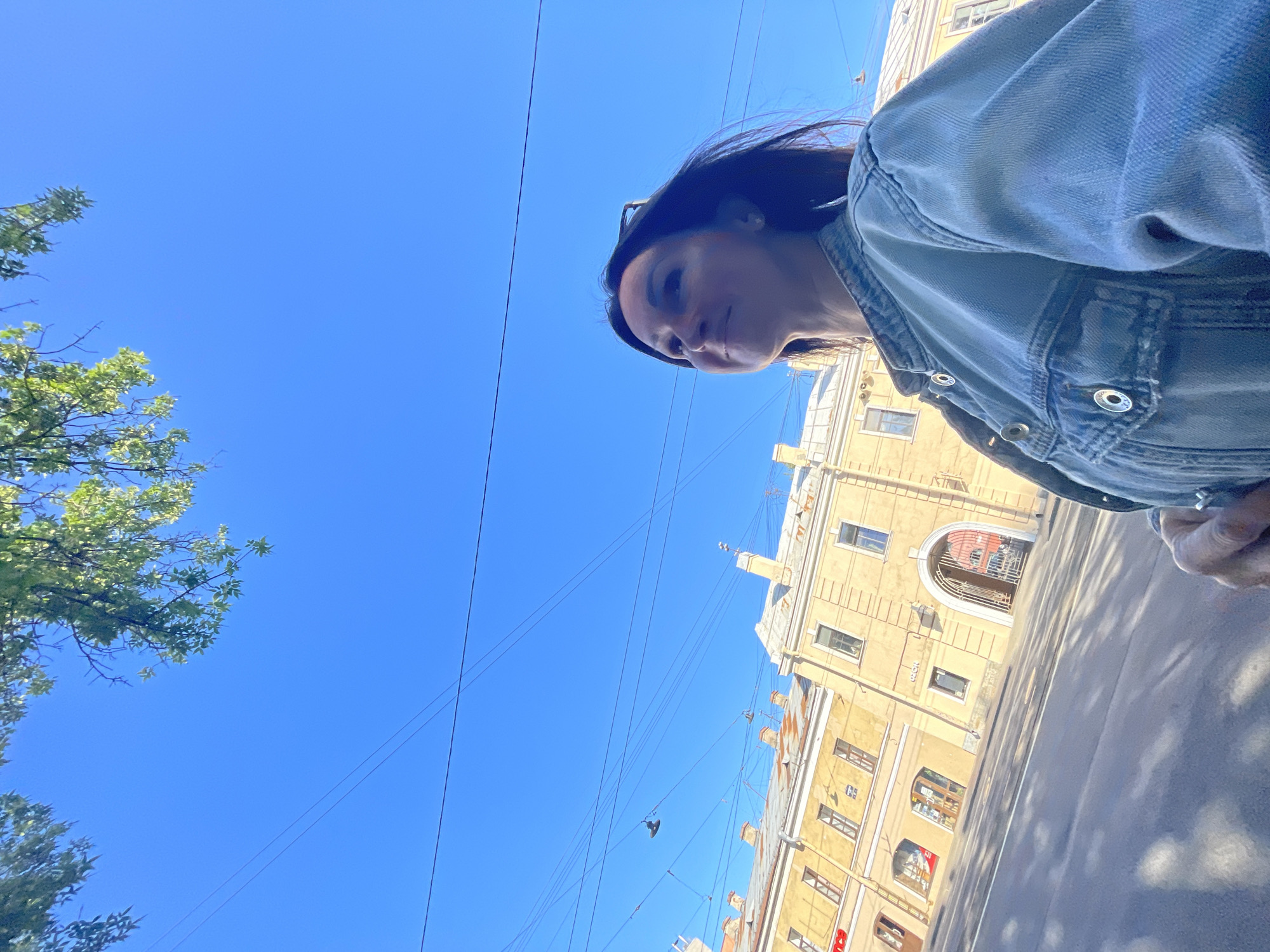 In Saint Petersburg, Russia, on a sunny day with a clear blue sky, a woman stands on a street. She is looking up and to her right, her dark hair is windswept. She is wearing a light blue denim jacket. Behind her, a light yellow building with a prominent archway is visible. The building has multiple windows and what appear to be shops on the ground floor. Overhead, tree branches with green leaves are visible against the sky, along with numerous overhead wires and a hanging lamp. The street in front of the building is paved and has shadows cast across it, suggesting a midday sun.