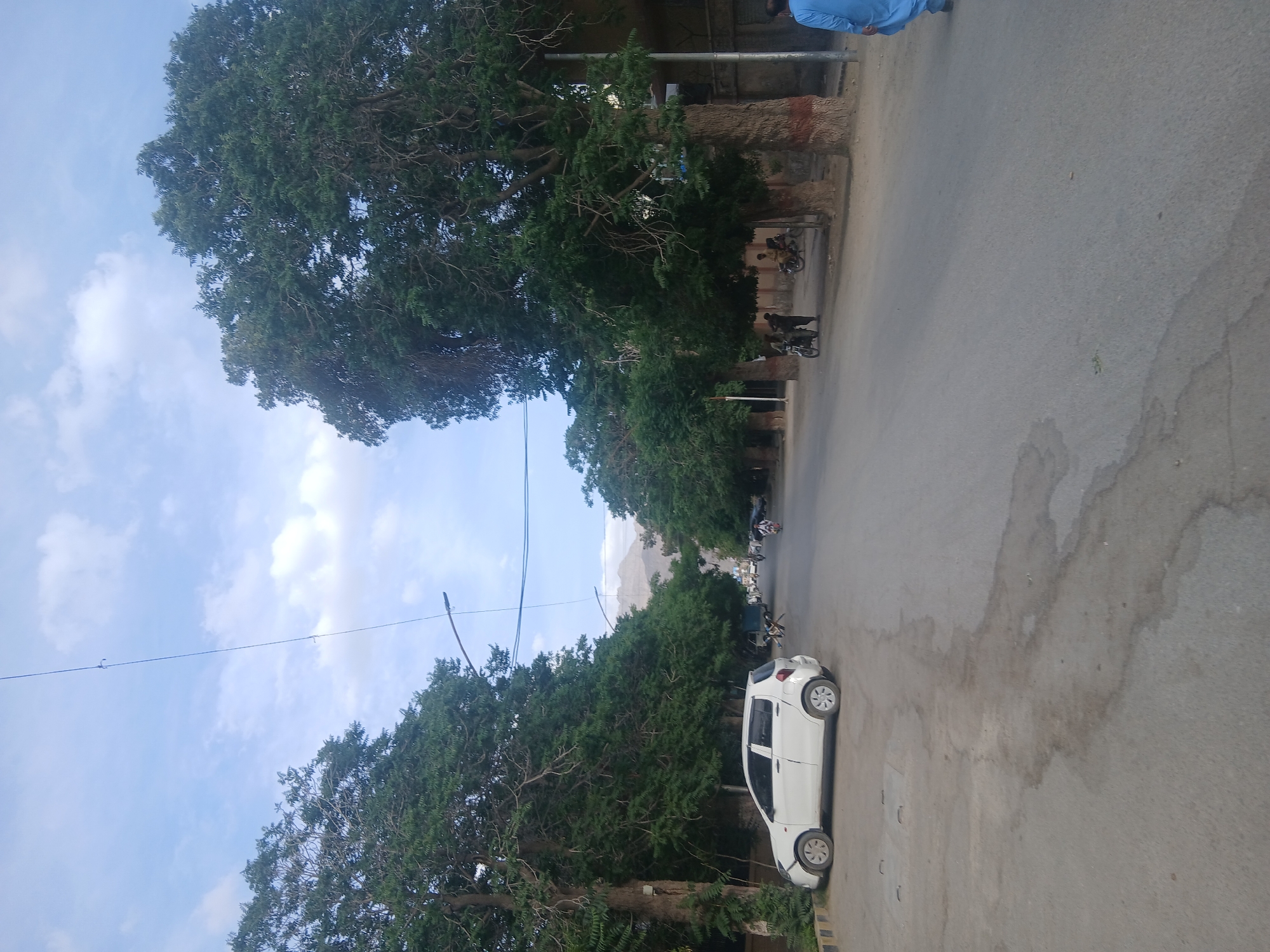 The image depicts a street scene in Quetta City Tehsil, Pakistan. A white car is parked on the right side of the road. Trees line the left side of the street, and a few people and motorcycles are visible further down the road. A mountain is partially visible in the distance. The sky is blue with some clouds. Power lines cross the sky. The road is paved.