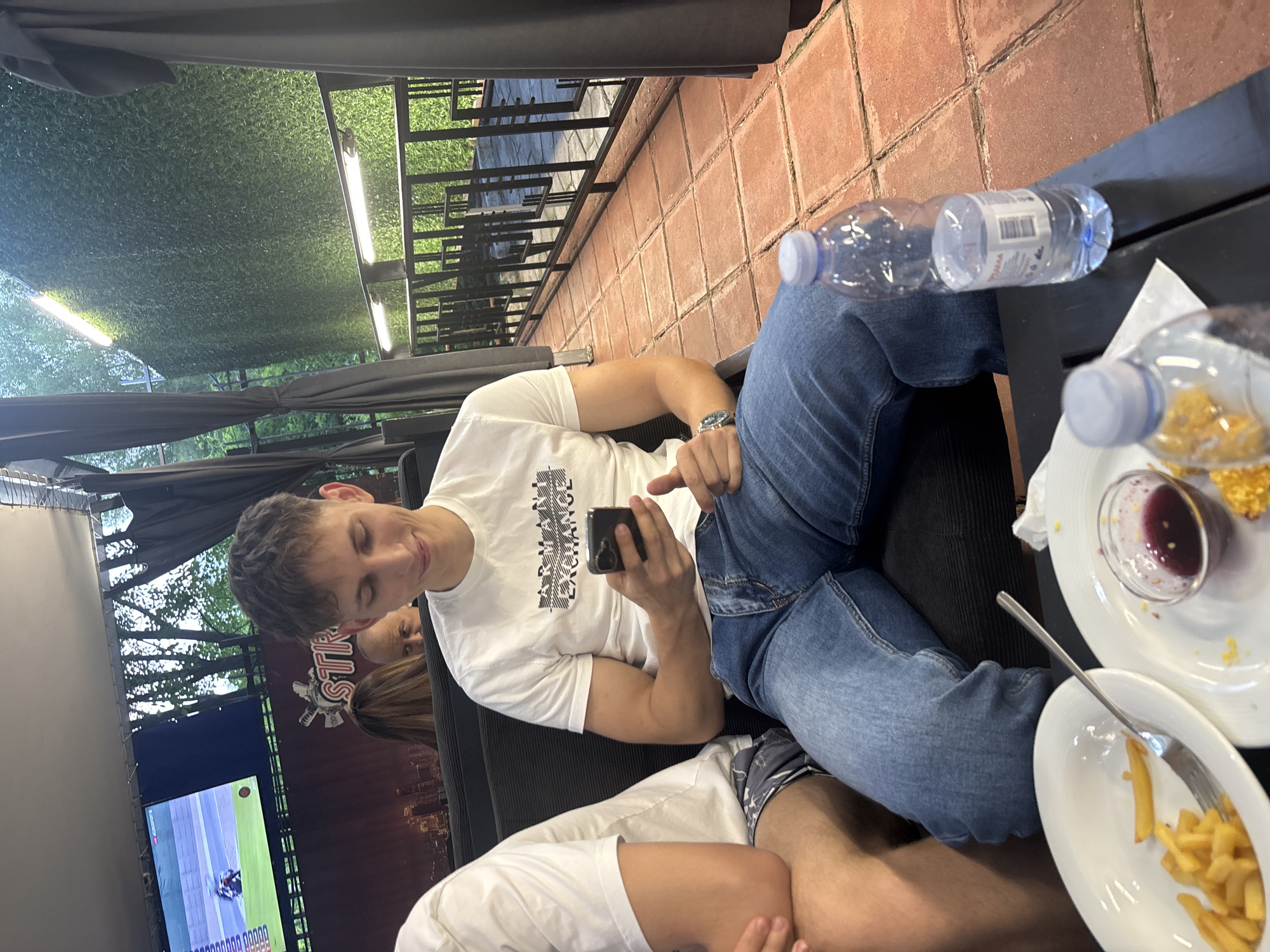 This image captures a semi-outdoor dining and entertainment area at Stryke Sports Bar & Bowling in Blagoevgrad, Bulgaria. A young man is seated centrally, wearing a white t-shirt with "ARMANI EXCHANGE" printed on it and blue jeans. He holds a dark-colored smartphone in both hands, looking down at the screen, with his left thumb positioned upwards. A silver wristwatch is visible on his left wrist.
To his left, another person is partially visible, showing a right arm in a white t-shirt and a right leg in patterned shorts. In the foreground, a dark table holds two white oval plates. One plate contains french fries and a silver fork. The other plate holds a small glass bowl with dark red sauce or jam and scattered yellow food crumbs. Two clear plastic water bottles are also on the table, one mostly full and the other partially consumed, resting on a white napkin.
In the background, a large screen displays a live motorcycle race, featuring a rider on a track with various graphical overlays. Below the screen, a sign with the partially visible word "STRYKE" and a logo is present. Behind the main subject, a woman with long brown hair is seated, and to her right, an older man's face is partially visible. The right side of the scene features a tall wall covered in artificial green foliage, illuminated by two horizontal strip lights. A dark metal railing with geometric patterns separates the seating area from an outer space where natural trees are visible. The floor is paved with reddish-brown square tiles, and dark grey curtains frame the outdoor setting.
