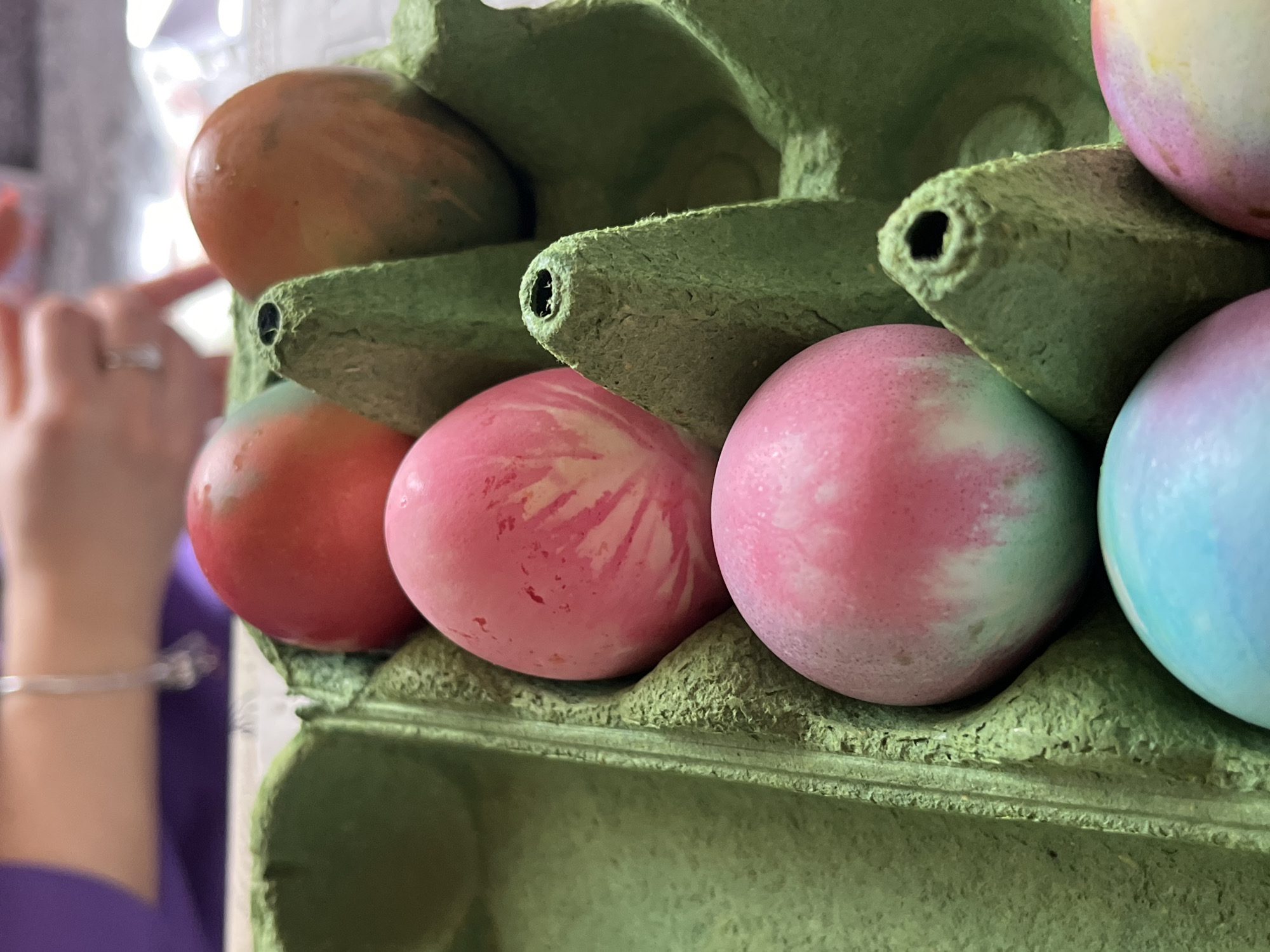 The image shows several Easter eggs, dyed in various pastel colors, sitting in a green cardboard egg carton. The eggs are primarily pink, red, and blue with a speckled or swirled pattern. A portion of a person's arm and hand is visible in the left side of the frame, suggesting someone is interacting with the carton. The background is blurry, obscuring further details of the environment. There is no indication or evidence to suggest the location is Kranevo, Bulgaria.