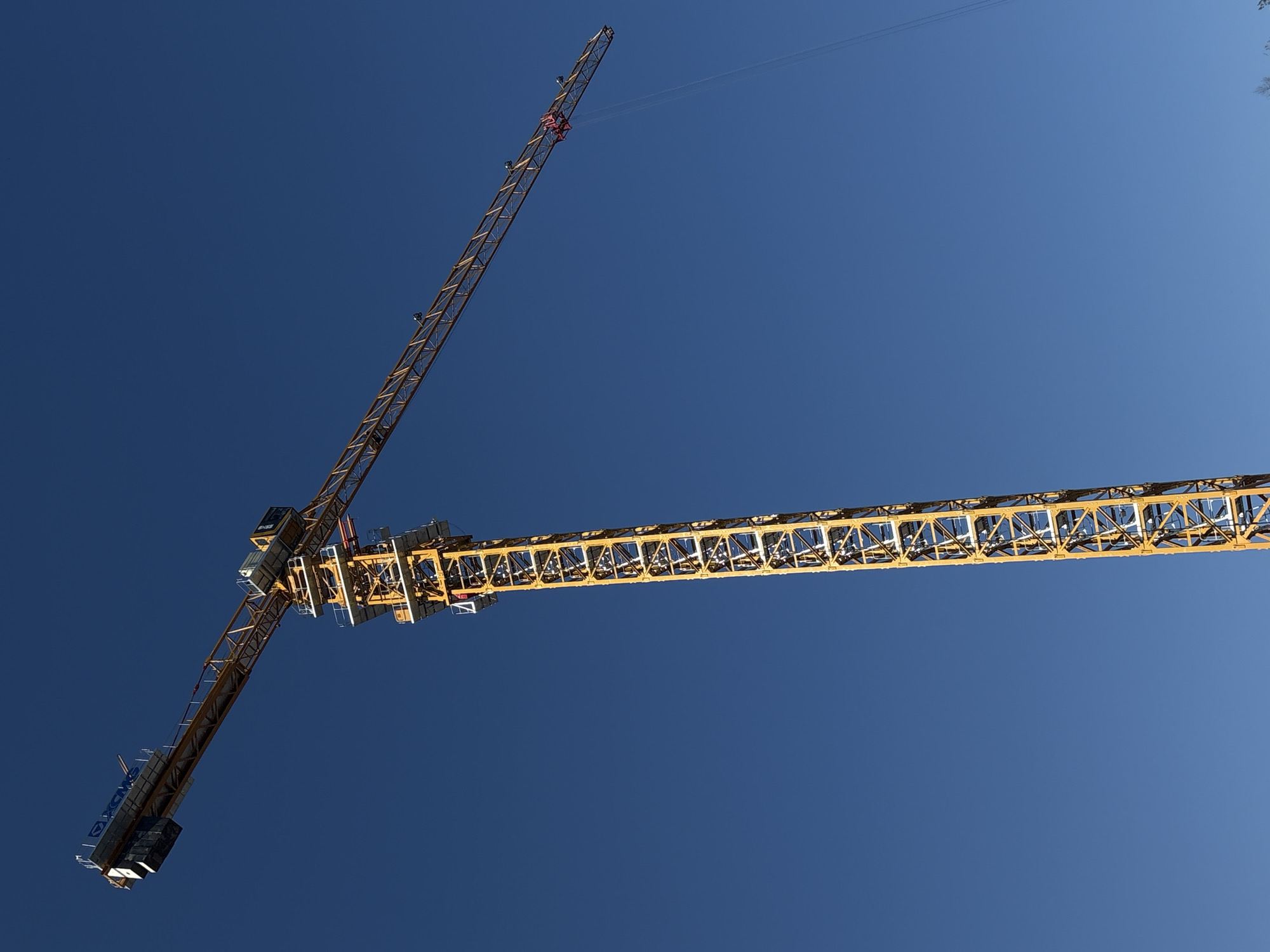 A low-angle shot of a yellow construction crane against a clear blue sky. The crane has two arms extending in different directions, with the main arm visible in the bottom right and the smaller arm pointing toward the upper left. The shot is taken from below, emphasizing the height and scale of the crane. The location is not discernable in the image.