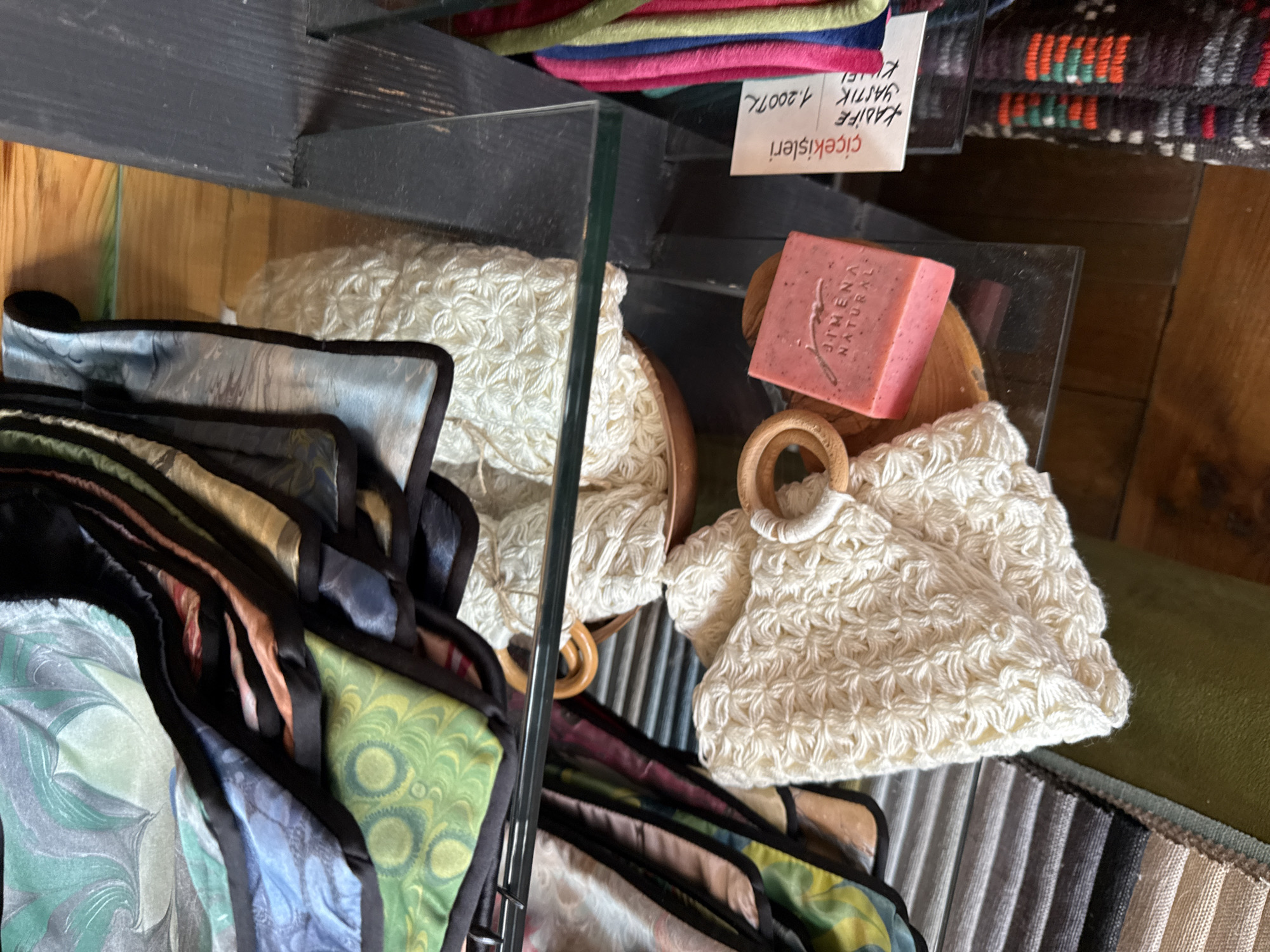 A close-up, eye-level shot reveals a retail display in Kadıköy, Turkey. The foreground showcases a woven, cream-colored object with a wooden ring, suggestive of a bath accessory or bag. To its right, a rectangular bar of pink soap, branded "JIMENA NATURAL," rests on a wooden surface. Adjacent to the soap is a white price tag with Turkish writing, including "çiçekışleri" and "1.200TL," indicating a price. Behind these items, on a glass shelf, are folded fabrics with intricate, marbled patterns in shades of green, blue, and earth tones. A dark wooden structure frames the right side of the image, and the background consists of rustic wooden planks. The lighting is soft, highlighting the textures and colors of the displayed merchandise.