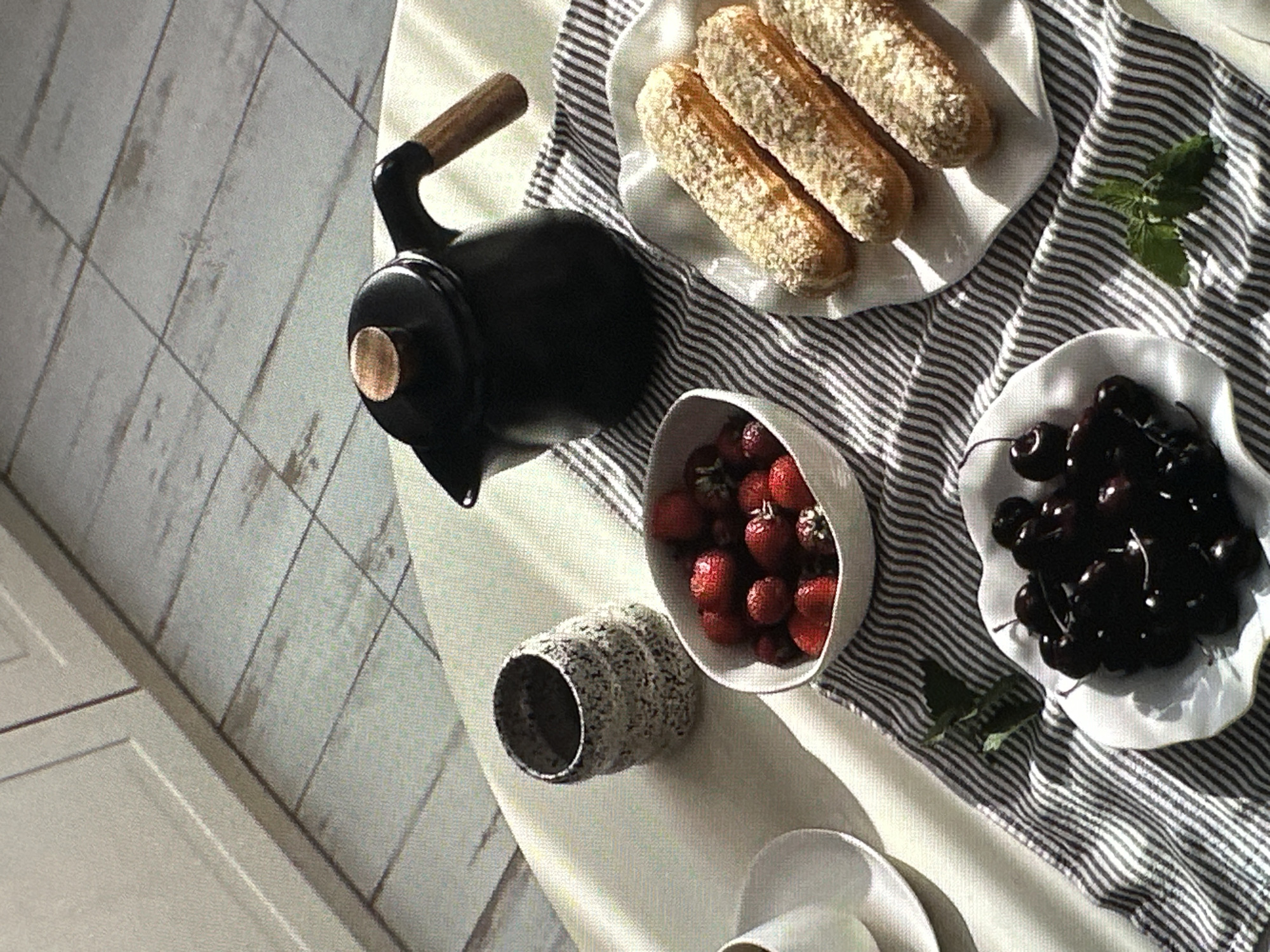 The image shows a still life arrangement on a striped fabric surface. A black kettle with a wooden handle is positioned near a white plate containing three pastries. Adjacent to the kettle is a white bowl filled with strawberries, and further along is another white bowl filled with dark cherries. A textured grey cup is also present near the strawberries. A white cup and saucer is visible in the lower part of the frame. Mint sprigs are scattered near the plates. The scene is illuminated by natural light.
There is no information present within the image to specify the location is Yerevan, Armenia.