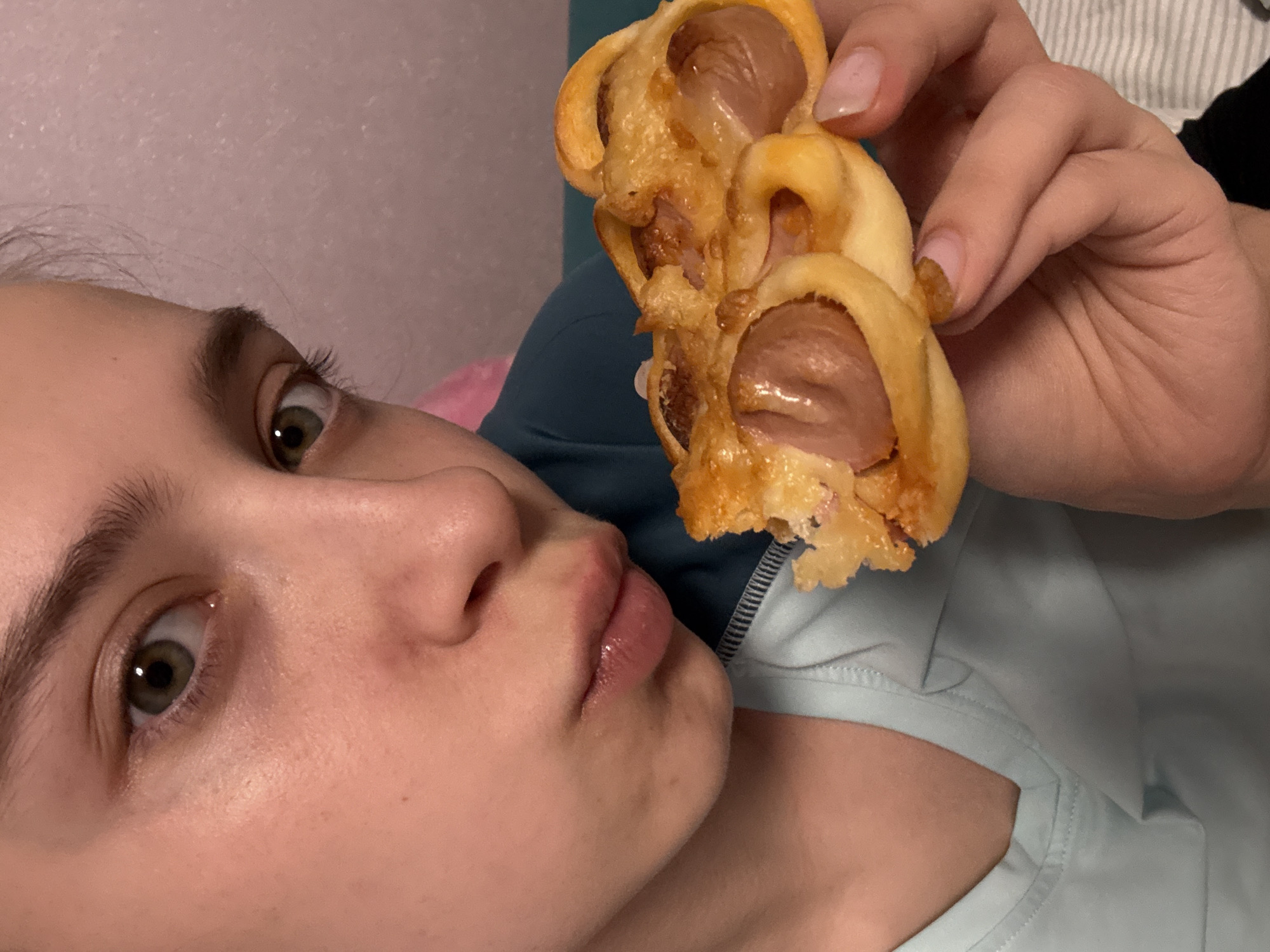 A young woman is shown in a close-up shot, holding a pastry filled with sausages in her right hand. The pastry has a golden-brown color, indicating it's likely baked. The woman is looking directly at the camera with a slightly surprised expression. Her eyes are wide and green. She has fair skin and her hair is light brown. She is wearing a light blue shirt or top with a visible neckline. The background is a blurry combination of pink and blue. There are no specific clues to pinpoint the location as Прикамский, Russia or any other specific city.