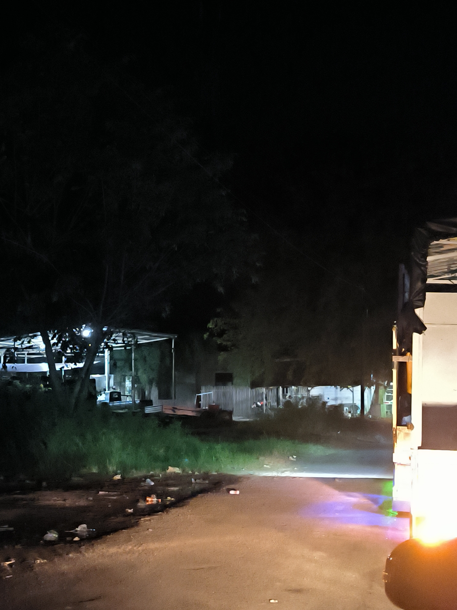 This night scene, set in Lhoknga, Indonesia, depicts a dimly lit outdoor area. In the foreground, a rough path or unpaved road is brightly illuminated by the headlight of a vehicle, a portion of which, appearing white or light-colored, is visible on the right side of the frame. The strong light casts distinct blue, purple, and green hues onto the ground directly in front of the vehicle. Scattered litter, including bottles and other debris, is visible on the ground.
A strip of green grass borders the path on the left. Beyond this, large, dark trees dominate the mid-ground and left side of the image, their branches silhouetted against the dark sky. Beneath the trees, several open-sided structures, possibly sheds or workshops with light-colored roofs and visible support beams, are faintly illuminated from within. A white fence or wall can be seen behind these structures on the right. The overall atmosphere suggests a utilitarian or industrial area after dark. No people or visible text are present.