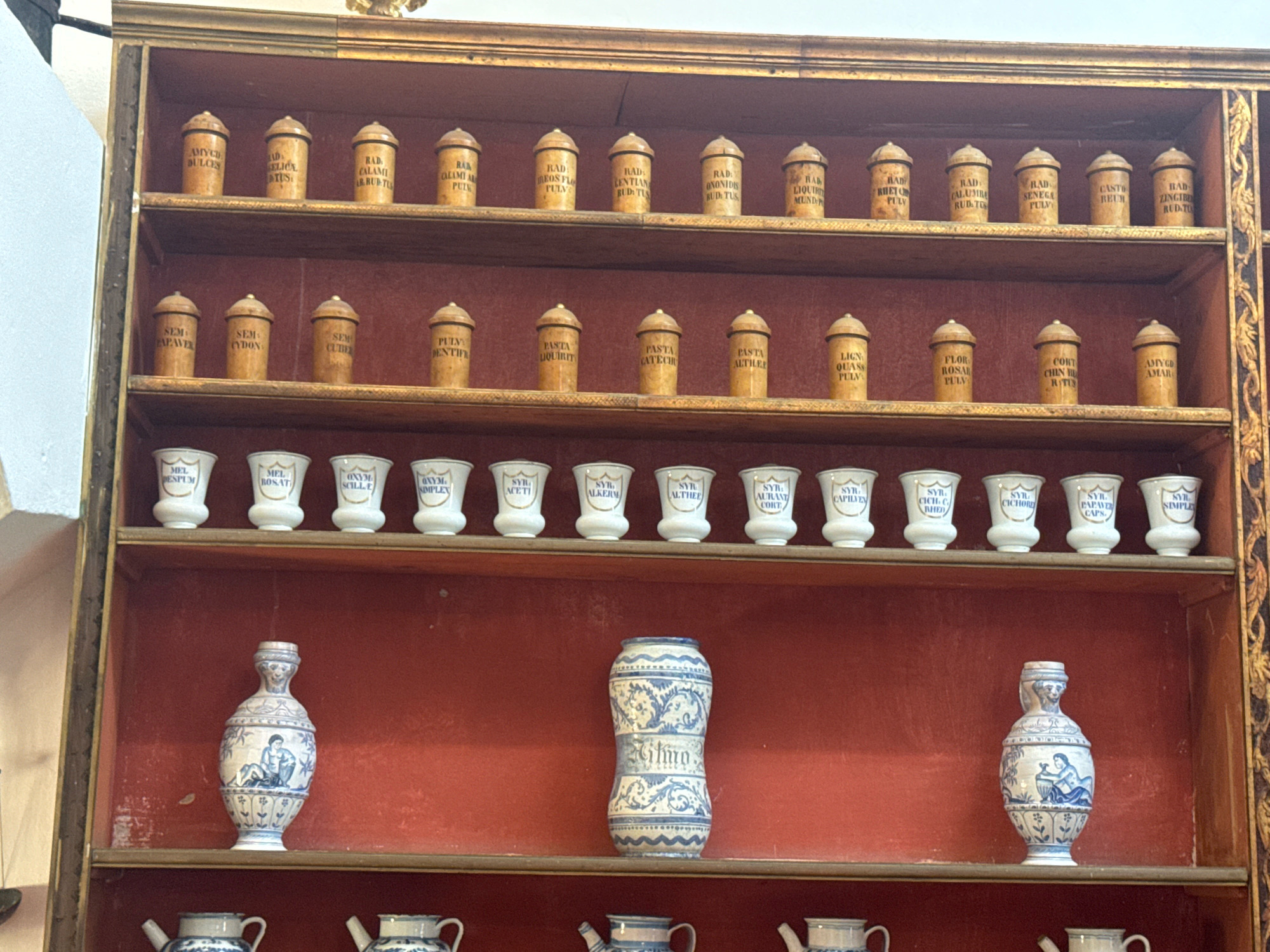 An antique apothecary shelf is displayed in a room in Dubrovnik, Croatia. The shelf is made of wood and has multiple levels. Each level is filled with various glass and ceramic jars and bottles. The jars on the top two shelves are small and made of wood, with labels indicating medicinal ingredients. The third shelf contains white ceramic jars with blue lettering. The bottom shelf features decorative ceramic vases and pitchers. The overall scene suggests a historical setting, possibly a museum or a preserved apothecary shop.