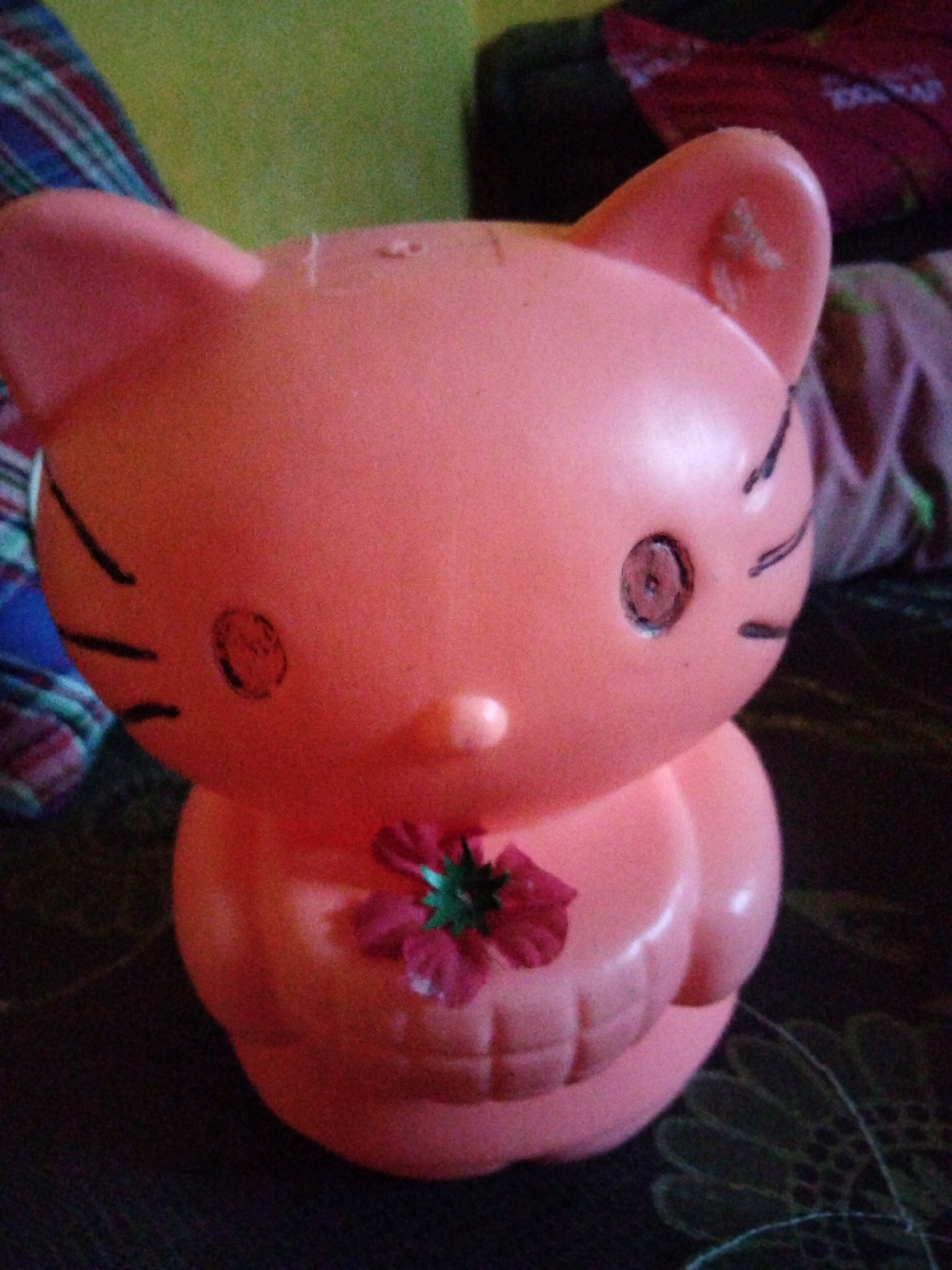A close-up, eye-level, medium shot captures a pink Hello Kitty-shaped piggy bank with a dark background and a green-yellow patterned cloth in the upper left corner. The piggy bank is the central focus, positioned slightly to the left of center. It has two pointed ears with faint black lines, black whiskers on either side of its face, and two black circular eyes with a faint inner detail, one appearing slightly smudged. A small pink flower with green accents is attached to the front of the piggy bank. The piggy bank is sitting on a dark, textured surface. The scene appears to be indoors, with no visible indication of the city of Payagaboh, Indonesia.