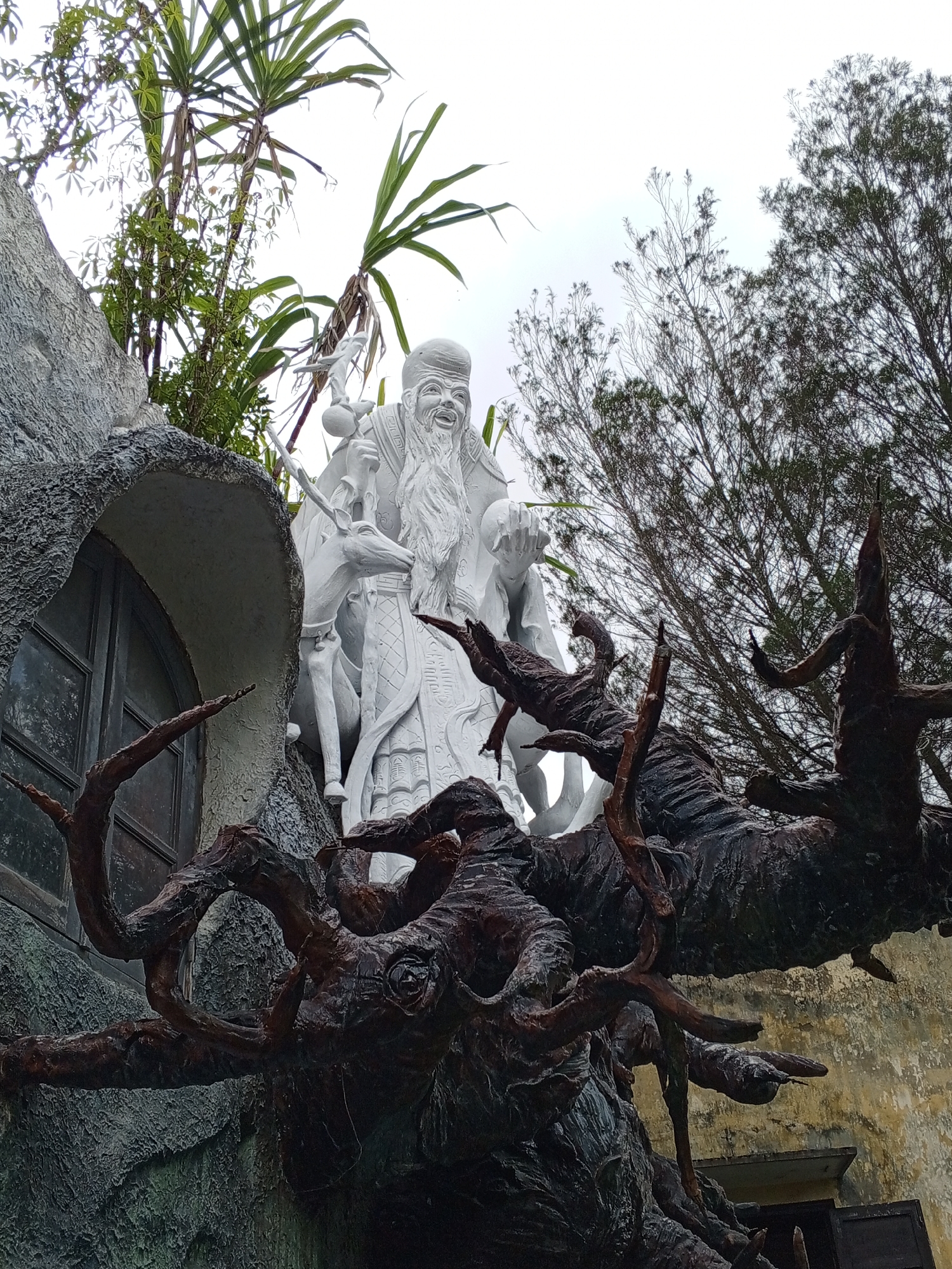The image captures a statue of an elderly man with a long white beard, wearing traditional robes and a hat. He is smiling and holding an orb in one hand and a staff in the other. Beside him stands a deer. The statue is made of white material and appears to be located outdoors, possibly as part of a garden or temple. In the foreground, there are dark, gnarled branches of a tree, adding a dramatic element to the scene. In the background, there are lush green plants and trees, as well as a cloudy sky. The architecture suggests an Asian setting, and the overall impression is one of a spiritual or mythical representation. This statue is located at the **Lam Ty Ni Pagoda** in Da Lat, Vietnam.