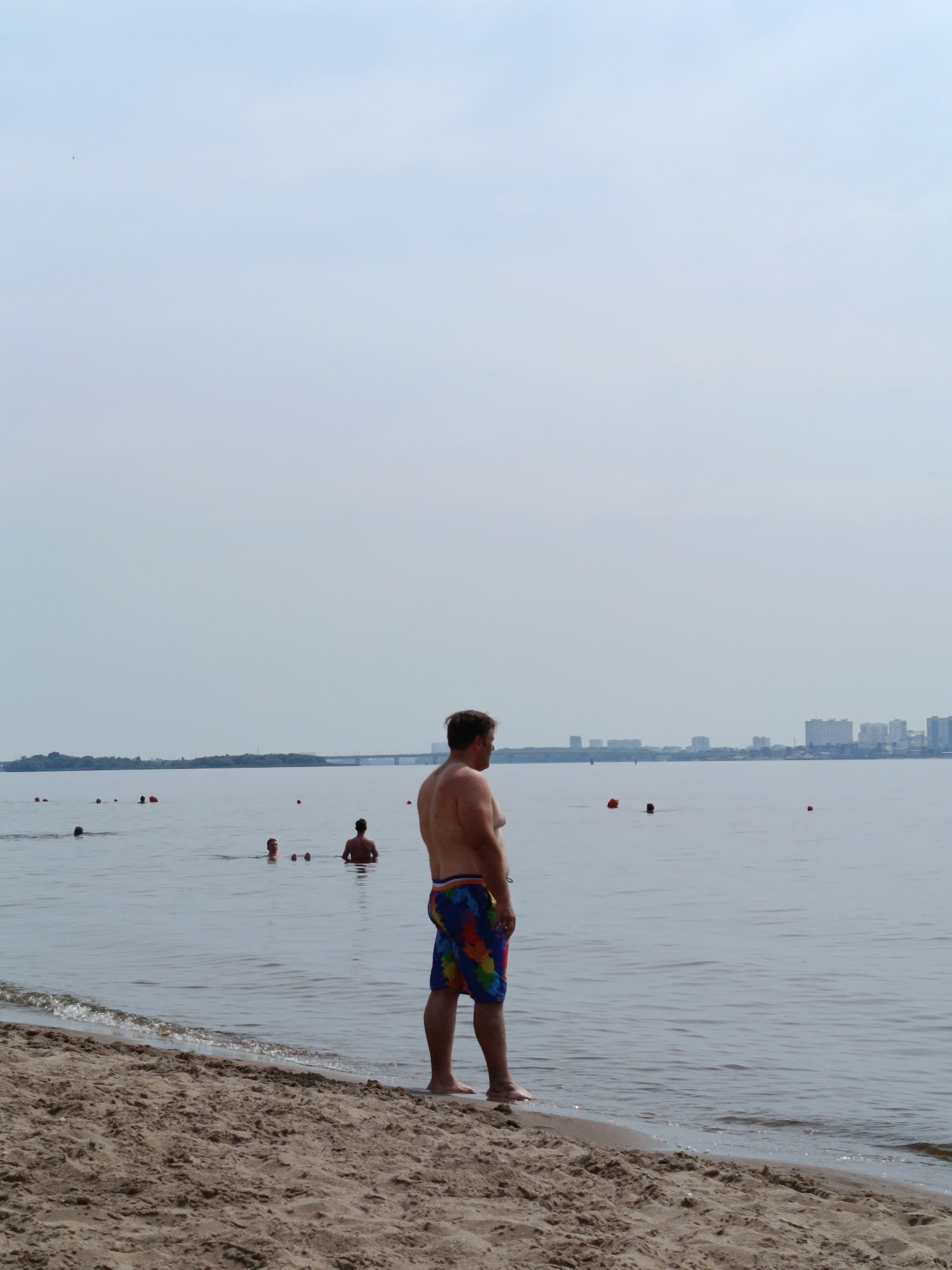 A man stands on the sandy shore of a body of water, facing away from the camera and towards the water. He is wearing colorful swim trunks. In the water, several other people are visible, some swimming and others standing. In the distance, a city skyline with tall buildings and a bridge can be seen across the water. The sky is overcast, and the water is calm. The scene depicts a typical day at a public beach.