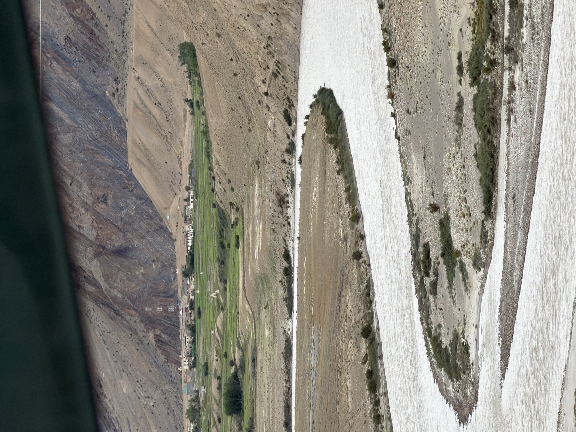 An aerial, wide-angle shot captures a wide, meandering river flowing through a dry, mountainous landscape. The river is a milky white color and takes up a significant portion of the foreground, with small islands and gravel bars visible within its flow. The banks of the river are lined with sparse, dry vegetation and rocky terrain.
In the middle ground, the landscape transitions to green agricultural fields, indicating a more fertile area. Several small, white-domed structures, likely stupas or chortens, are scattered amongst the fields. A small village with white-washed buildings and a prominent cell tower is nestled at the base of the mountains.
The background is dominated by steep, rocky mountains with visible layers of sediment, suggesting a geologically complex area. The sky is overcast and a dark green band is visible across the top of the frame, likely the edge of a banner or prayer flag. The image depicts a remote, high-altitude settlement in Zangla, India, characterized by its stark natural beauty and the presence of human habitation adapted to the harsh environment. There are no people or animals visible in the image.