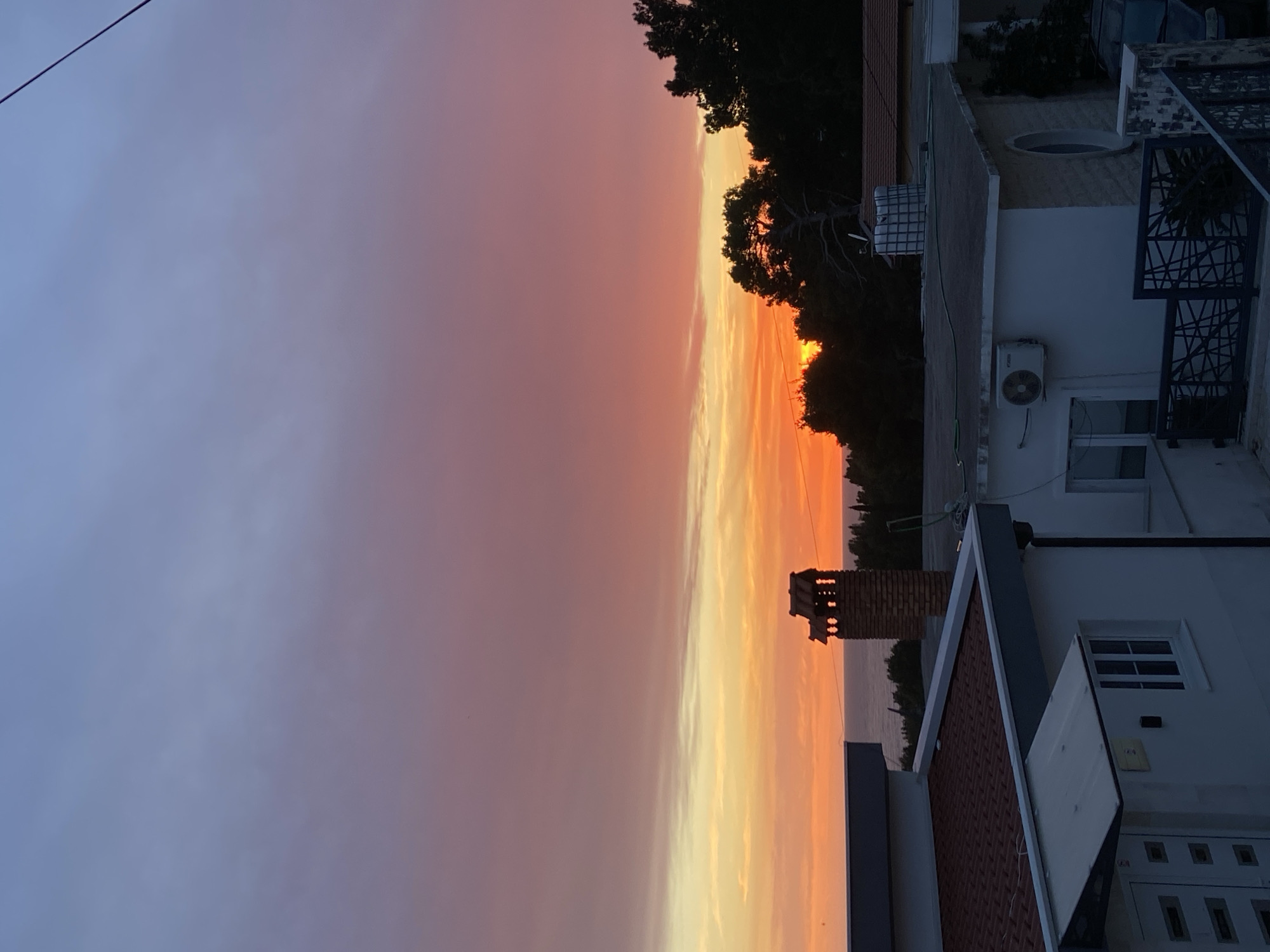 The image captures a sunset in Šušanj, Montenegro, with the horizon showing a blend of yellow, orange, and red colors fading into a purple-gray sky. In the foreground, there are rooftops of buildings, some with red tiles, and silhouettes of trees. A brick chimney is visible, alongside details like an air conditioning unit, windows, and decorative metalwork on the buildings. A power line is visible in the upper left corner. The scene portrays a quiet, residential area under a colorful sunset sky.