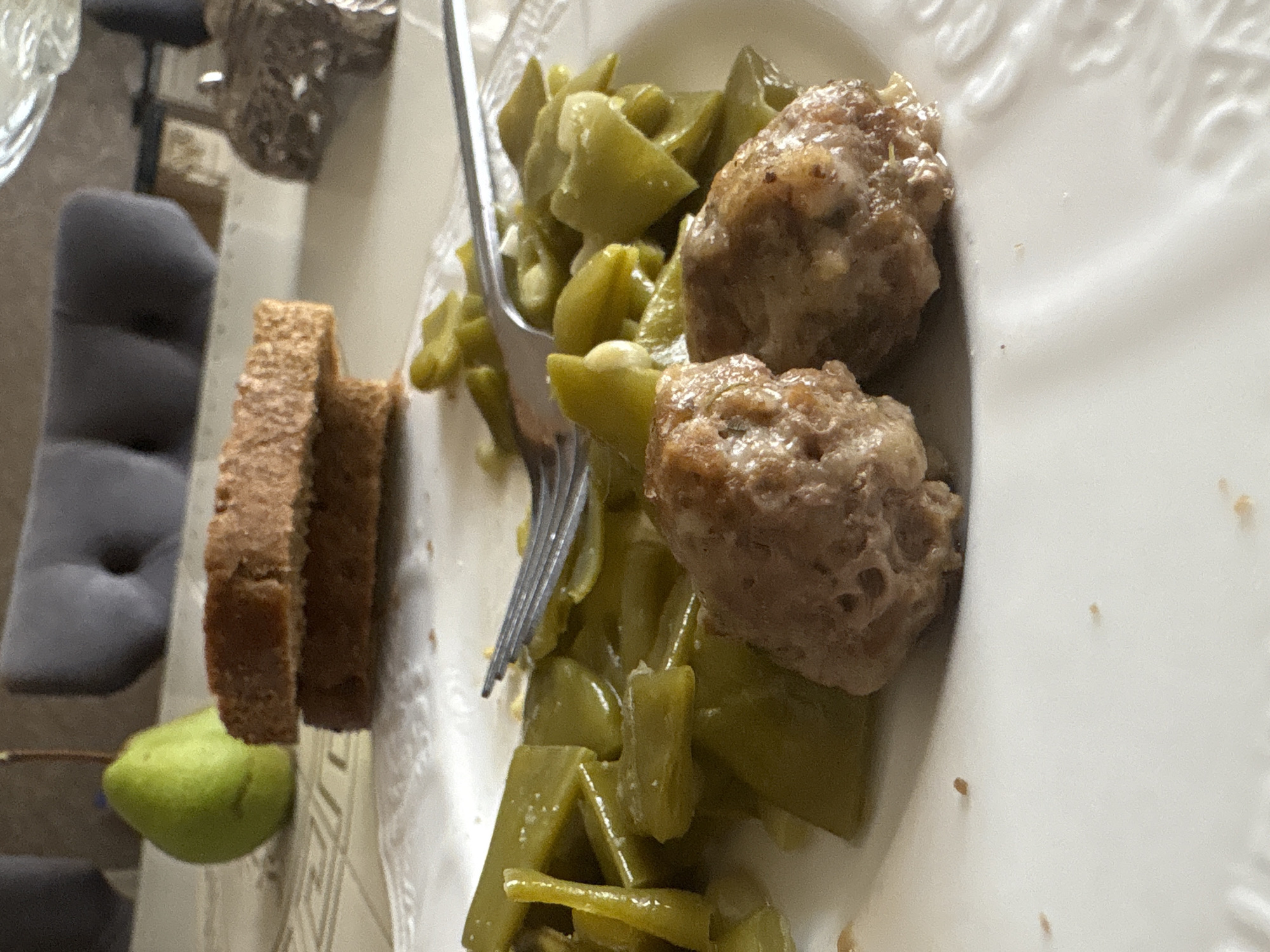 A plate of food, including green beans and meatballs, sits on a table. In the background, a green pear and two slices of bread are visible. The scene appears to be indoors, possibly in a home or restaurant. The image is taken from a close-up perspective, focusing on the food.