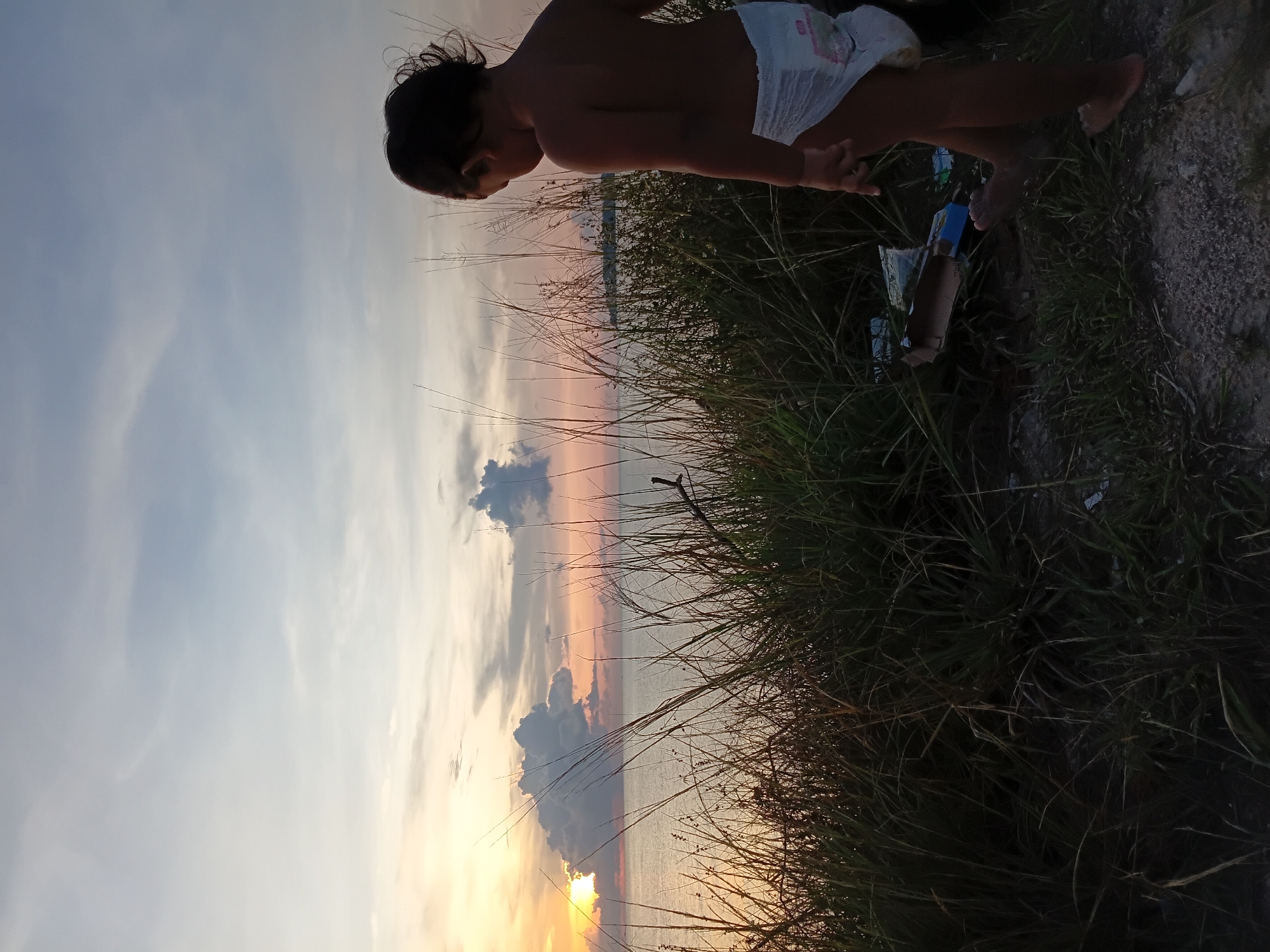 A medium shot of a child in a diaper on grass near debris at Baan Wok Tum, Thailand, with a sunset view. The child stands on the right of the frame, close to discarded items on the ground. Tall grass covers a substantial portion of the bottom half of the image. The sky is visible as the backdrop, indicating a sunset. The horizon shows the ocean.