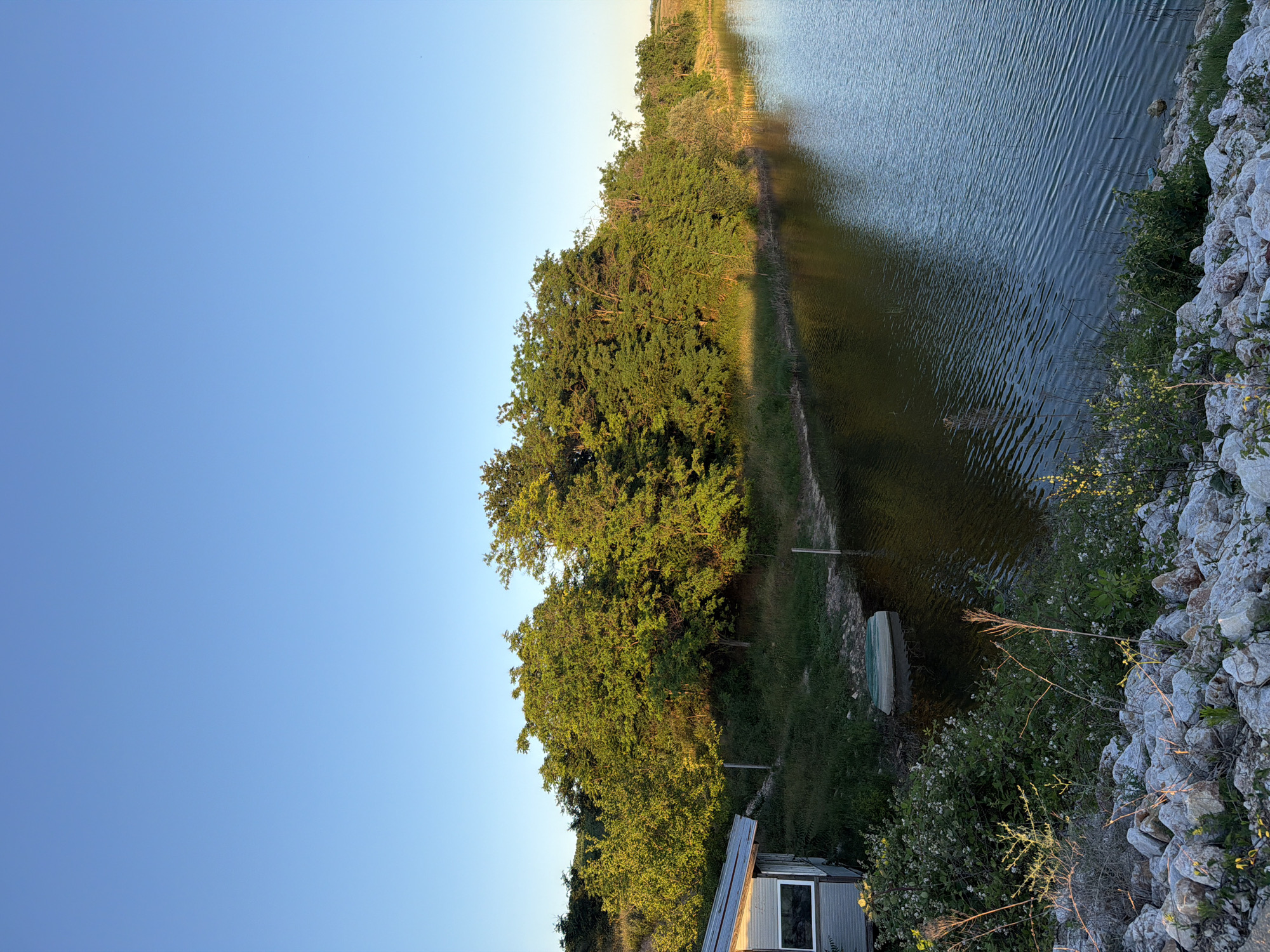 The image depicts a serene lakeside scene with a clear blue sky overhead. In the foreground, a rocky embankment lines the water's edge, interspersed with sparse vegetation. A small, light-colored structure with a gray roof and a window is partially visible on the left side of the frame. Adjacent to the structure, on the grassy bank, a green boat rests in shallow water. A line of lush green trees and foliage stretches along the opposite bank of the lake, with the setting sun casting a warm glow on the leaves. The water's surface is rippled, reflecting the surrounding greenery and the clear sky. The overall atmosphere is peaceful and natural, suggesting a remote or rural location. The image does not contain any people or evidence of human activity beyond the structure and the boat.
