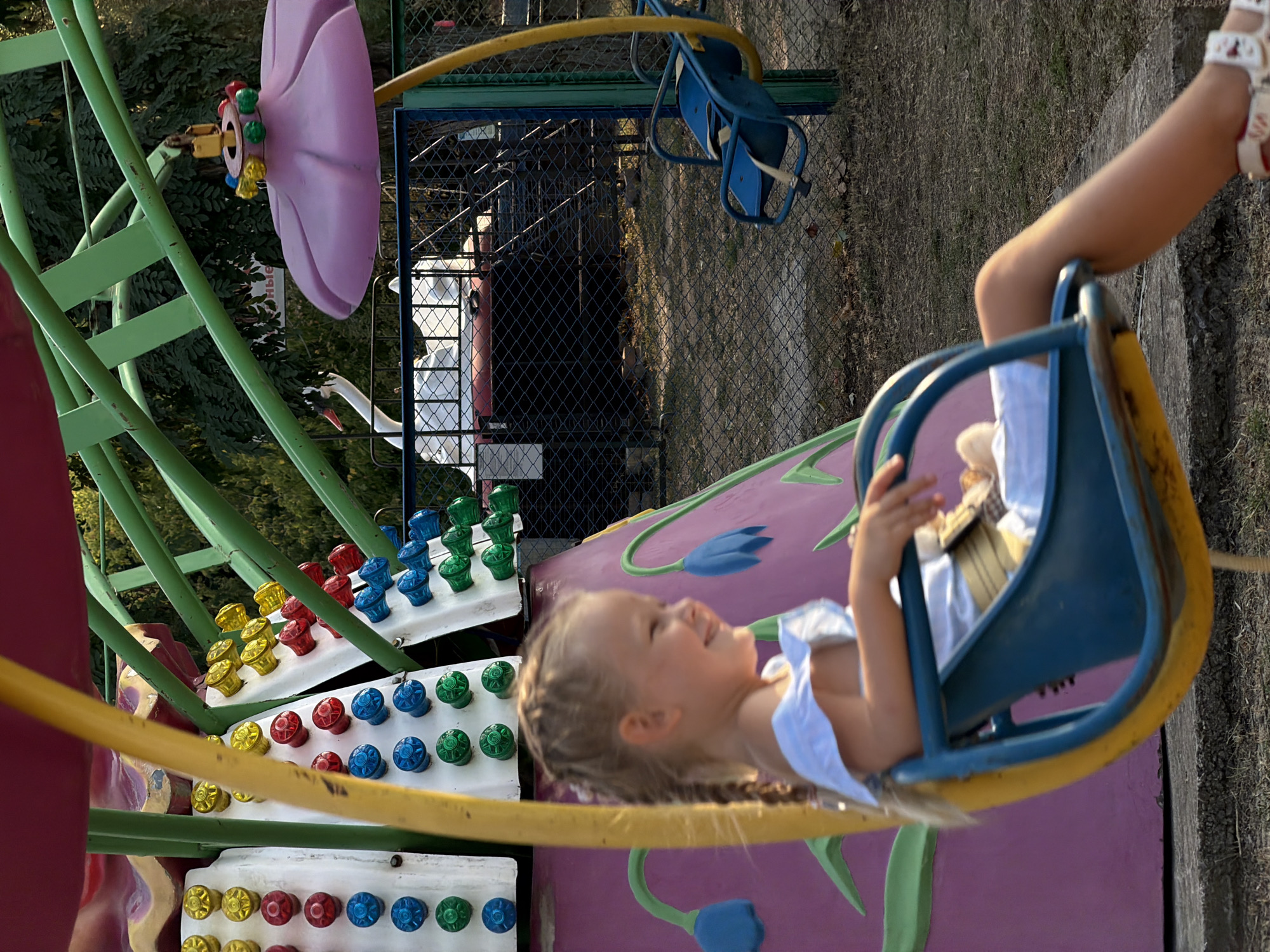 In Kamyshin, Russia, a young girl with blonde braided hair rides a swing at an amusement park. She is wearing a white off-the-shoulder top and white shoes. The swing is blue with yellow supports. In the background, there are colorful lights on a ride, a pink umbrella-shaped decoration, and a fence. Trees and foliage are visible behind the fence.