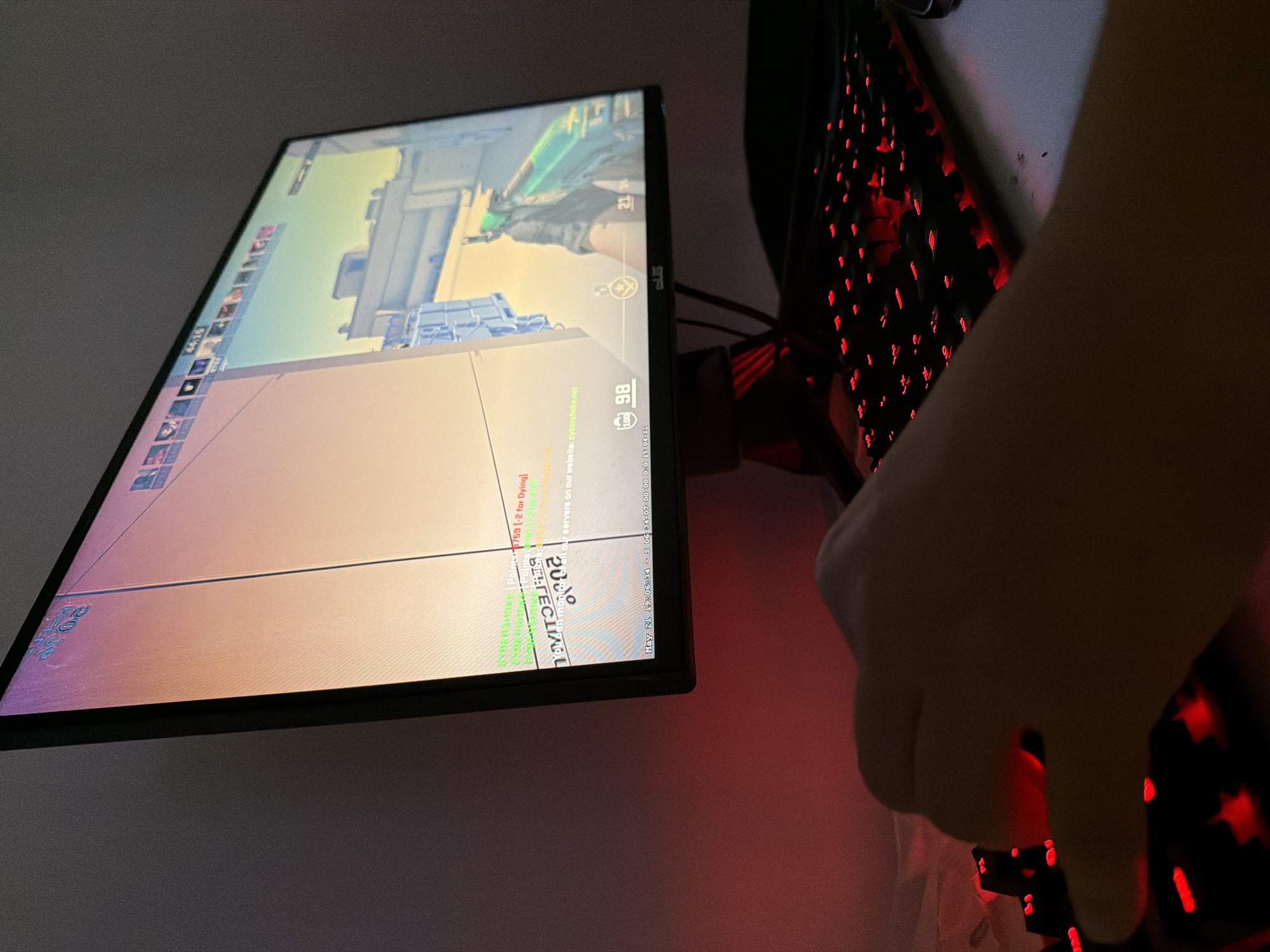 The image depicts a person playing a first-person shooter video game, likely on a computer. The monitor displays the game interface with the character's health and other in-game details visible. The person's hand is visible on a gaming mouse, and a red-lit gaming keyboard is in the background. The game itself appears to be Counter-Strike or a similar tactical shooter. OCR revealed '2030' 'cyberhoke' 'Dying', 'May 21'.