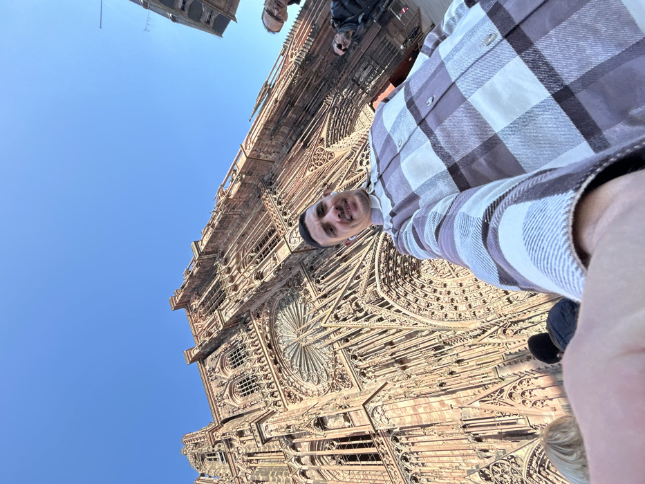 A low-angle selfie captures a male individual positioned in the foreground against the backdrop of the Strasbourg Cathedral in Strasbourg, France. The subject, a male in his 20s or 30s with short dark hair, a beard, and white wireless earbuds, smiles subtly while wearing a light purple and white plaid long-sleeved shirt. His right arm, extended towards the camera, is visible in the lower right. The expansive, intricate Gothic facade of the Strasbourg Cathedral, constructed from reddish-pink sandstone, dominates the background, showcasing its elaborate arches, numerous windows, and a prominent rose window above the subject's head. The sky is clear and brilliant blue. Several other individuals are present in the deeper background: two partially visible heads to the left of the main subject, and two figures to the right, one of whom wears glasses. The scene suggests a sunny public setting in front of the cathedral.