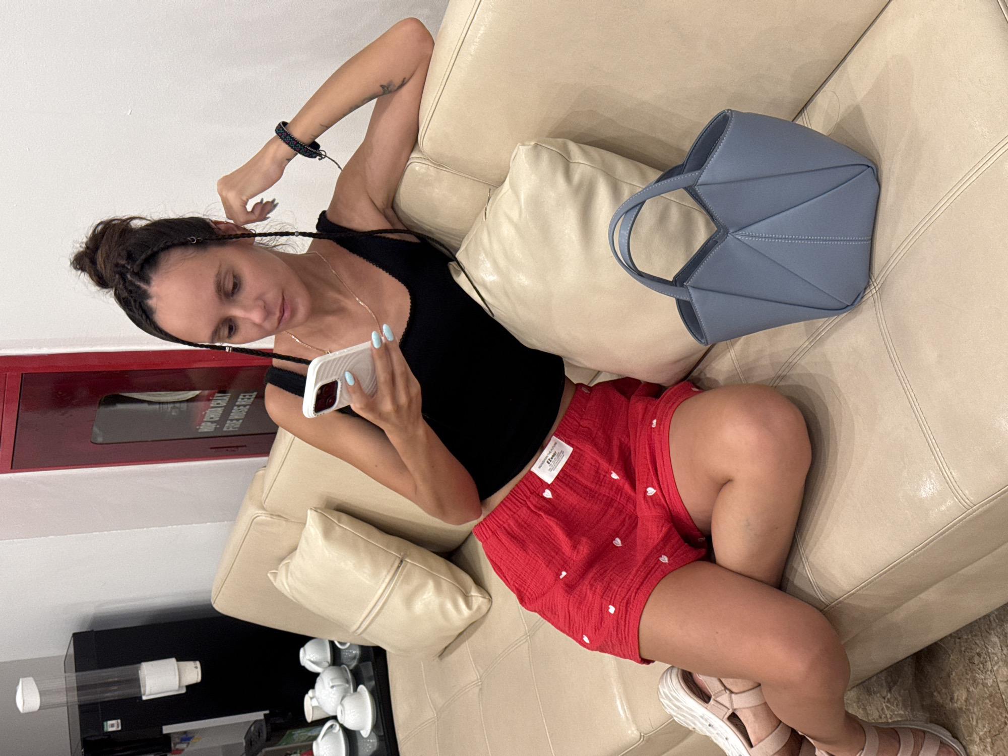 In Hà Nội, Vietnam, a woman with dark hair in a bun and braids sits on a beige leather sofa, holding a smartphone. She wears a black tank top and red shorts with white heart patterns. She has light blue nail polish. To her right is a light blue origami-style handbag. Behind her, a black dispenser with cups is visible on the left, and a red fire hose reel is on the right. There are several white teacups and saucers on a table in the background.