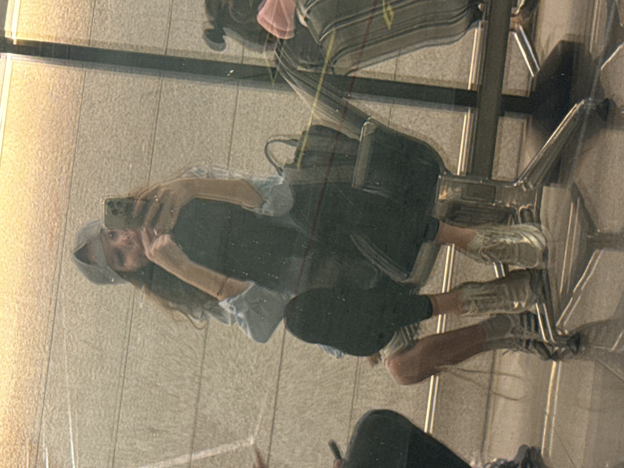 The image displays a mirror selfie taken indoors, likely in a public waiting area such as an airport, given the context of Vnukovo, Russia. The primary subject is a young woman, seen in her reflection, who is seated on a modern chair with a metallic frame and a dark upholstered seat.
The woman has long, dark hair and is wearing a light-colored, possibly light blue or grey, button-up shirt or jacket over a darker top. She is also dressed in dark pants or a skirt and light-colored sneakers with visible laces. A grey hood or cap covers her head. She is holding a smartphone, identifiable by its multiple camera lenses on the back, in her right hand, aiming it to capture her reflection. Her left arm is bent, with her hand near her chest. She is seated with her left leg bent and foot resting on the chair's base, while her right leg is extended slightly.
A dark bag is visible, either hanging on the chair or placed next to it. The background reflected in the mirror shows a light-colored, textured surface, resembling tiled flooring or a wall. A prominent dark horizontal line runs across the reflection, possibly a structural element of the room or a seam in the mirror. The scene is illuminated by artificial indoor lighting, which creates highlights on the metallic components of the chair. Faint reflections, possibly of another person's lower body or additional furniture, are present in the lower right and upper left areas of the mirror. There is no visible text in the image.