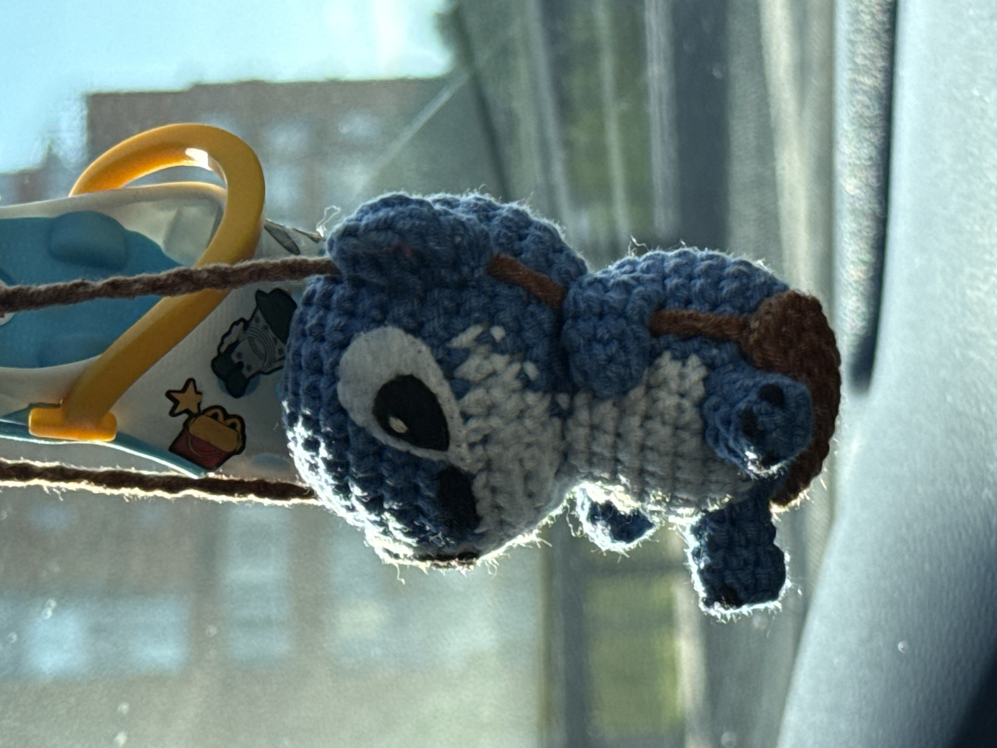 An outdoor medium shot captures a crocheted doll, resembling the character Stitch from Disney's Lilo & Stitch, hanging from a car's rearview mirror. The doll is predominantly blue with white accents and has black eyes and small black paw prints. It's suspended by a brown rope from a yellow plastic car accessory which is attached to a patterned blue and white fabric. The background is blurred but shows what appears to be a building with windows, suggesting an urban environment. The lighting suggests it's daytime. The location is unspecified and the image does not provide any details that would allow for identification of the city of Kyiv, Ukraine.