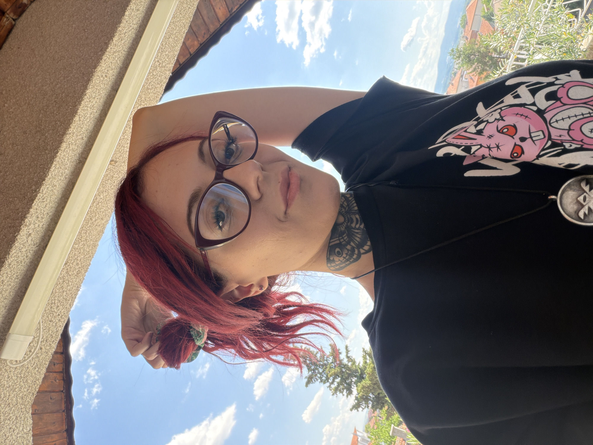 The media file shows a young woman with red hair taking a selfie outdoors. She is wearing glasses and a black t-shirt with a graphic print of a pink cartoon rabbit skeleton. A pendant necklace hangs around her neck. Her left arm is raised behind her head, and her right hand is holding her hair. She has tattoos on her neck. The background consists of a blue sky with white clouds, a balcony railing, and a glimpse of buildings and trees in the distance. The lighting suggests it is daytime, likely late morning or afternoon, with bright sunlight. The overall mood appears casual and relaxed. The location context is Svilengrad, Bulgaria.