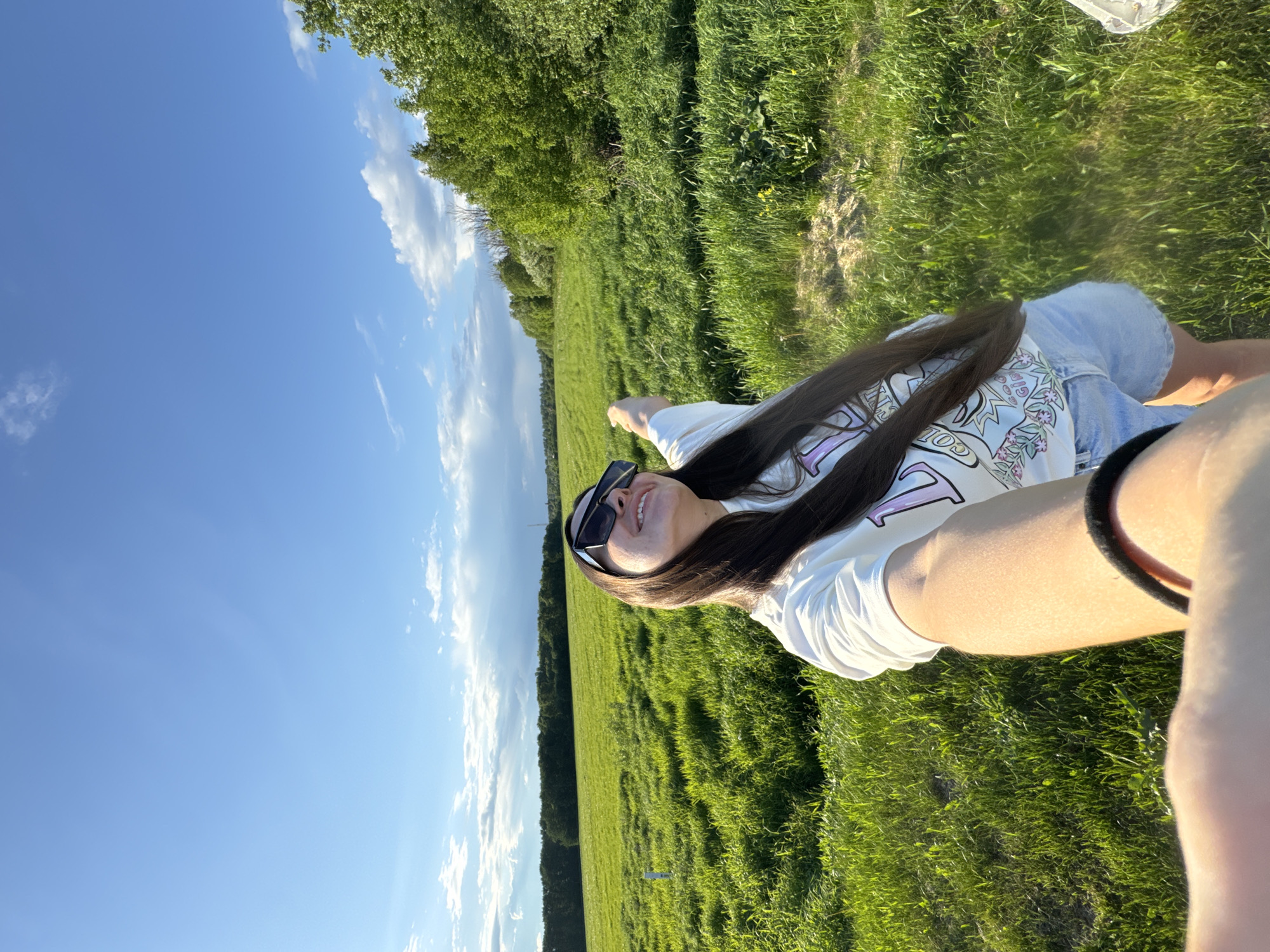 A medium shot shows a woman wearing sunglasses and a light-colored t-shirt lying on a field of green grass under a blue sky with some clouds. Part of a forest is visible at the edge of the field. The photo appears to be taken in a rural area, although it is not possible to confirm it is in the city of Bolshiye Vyazyomy, Russia based on the image. The camera angle is tilted.