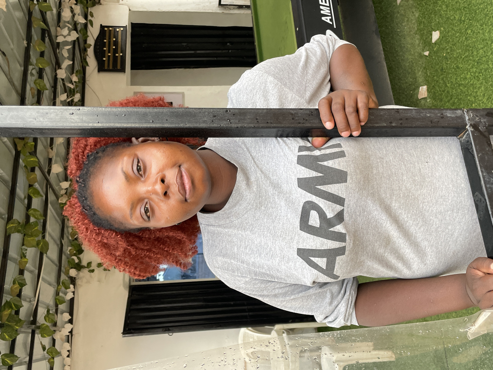 A woman with red curly hair is seen from the chest up, looking at the camera, while leaning on a black metal frame. She is wearing a gray "ARMY" t-shirt. The location appears to be outdoors, with vegetation visible in the background and to the left of the subject. A sign and a door are visible behind the woman. The scene is taken in daylight. The city of Jos, Nigeria, is not discernible from the image.