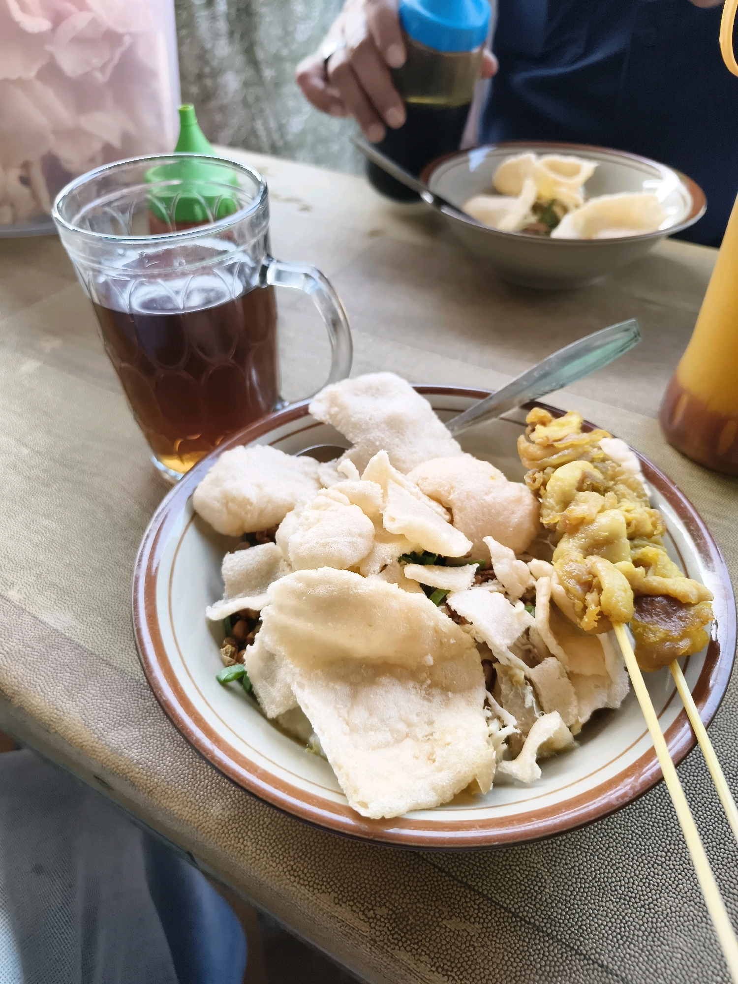 A close-up overhead shot shows a ceramic bowl of food on a patterned textured surface, likely a table, in a casual dining setting in Salira, Indonesia. The bowl contains a mixture of shredded chicken or meat, topped with numerous white, irregular-shaped crackers. Two yellow skewers with pieces of meat are positioned on the right side of the bowl. To the left of the bowl is a clear glass mug filled with a dark brown liquid, presumably tea. In the background, out of focus, a hand is holding a small bottle with a blue cap and a dark liquid, while another person's arm and shoulder are visible wearing a dark blue shirt. A tall, clear plastic container with a pinkish interior and a green spout is to the left of the mug, and a yellow bottle is partially visible on the far right.