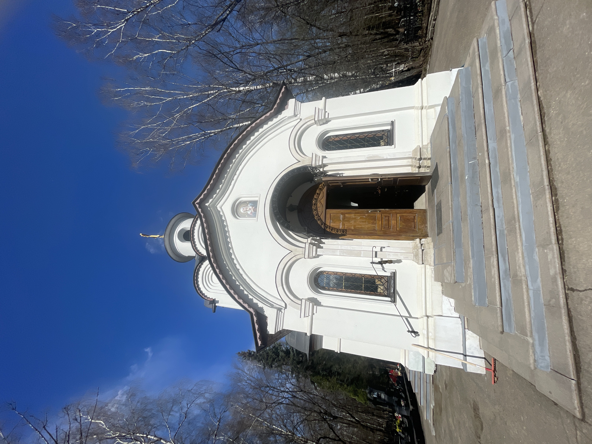 The image shows the Chapel of the Holy Martyr Uar in the Danilov Cemetery in Moscow, Russia. The chapel is a small, white building with a curved roofline and a small dome topped with a golden cross. There are arched windows on either side of the entrance, and the wooden doors are open. A set of stone steps leads up to the entrance. The sky is a clear, bright blue, and bare trees are visible around the building. There are headstones in the background.