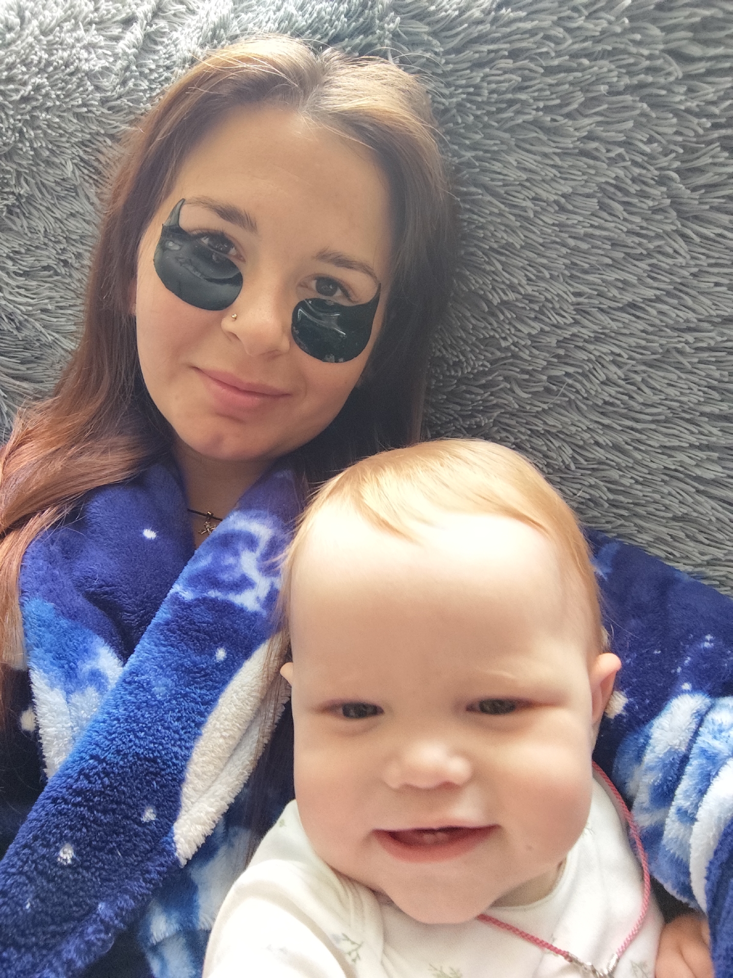 A medium shot shows a woman with long auburn hair, wearing black under-eye masks and a blue and white galaxy-patterned robe, embracing a baby with blonde hair who is smiling. They are lying down on a gray, fluffy blanket. The background is out of focus and consists of the gray blanket. The scene appears to be indoors, possibly in a bedroom or living room. The lighting is soft and natural. There is no indication of the city of Фоки, Russia.