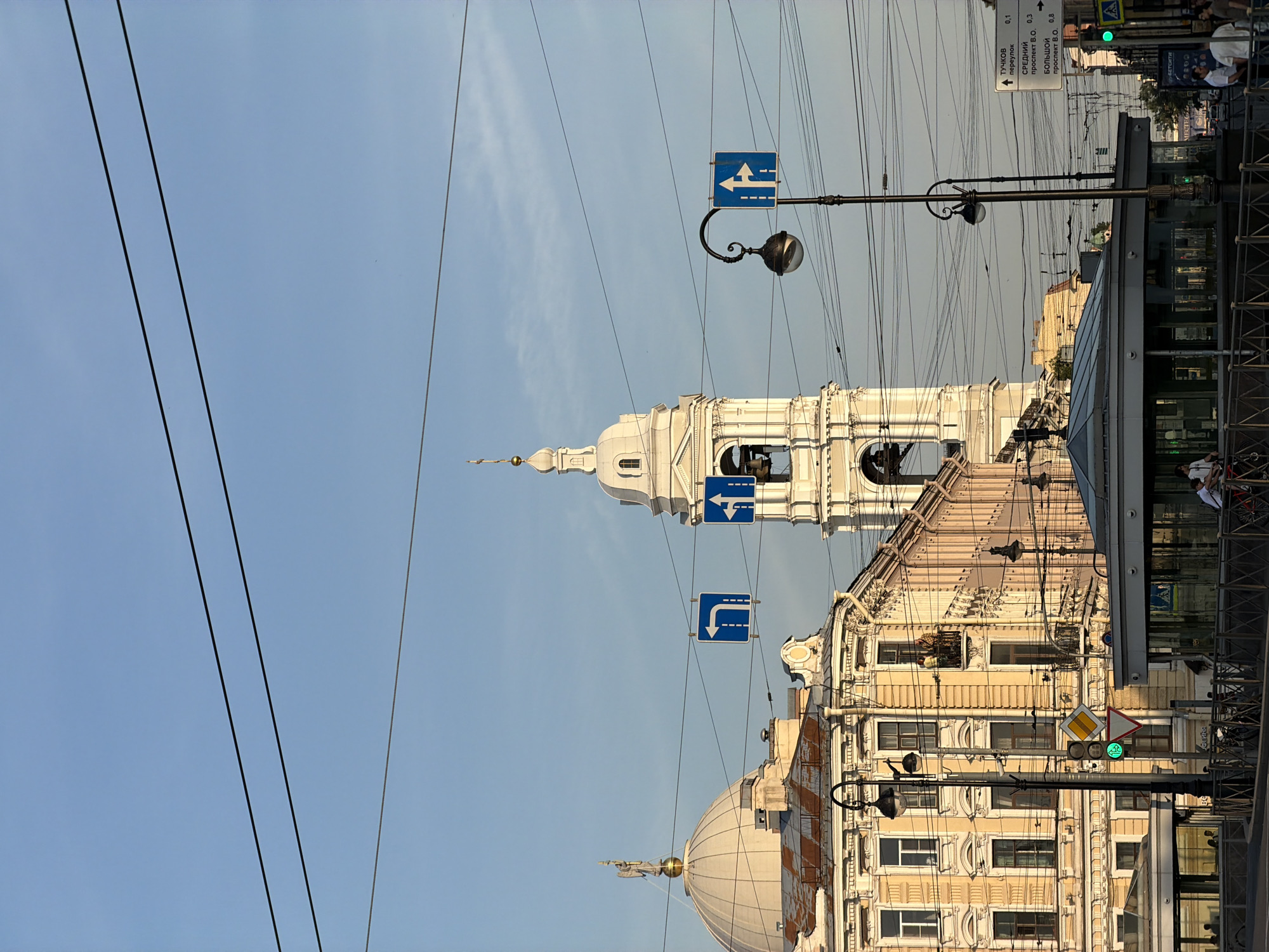 This is a street-level, eye-level photograph taken in Saint Petersburg, Russia. The scene is dominated by the architecture of the city, with a prominent bell tower of a church or cathedral on the right and a domed building on the left. Yellow and pink buildings with ornate details line the street. Overhead, a complex network of electrical wires crisscrosses the sky, characteristic of urban environments.
Several traffic signs are visible: blue directional signs indicate left turns and straight paths, while a green traffic light is illuminated. A street lamp with a classic design stands near the center of the frame. In the foreground, a modern glass-enclosed structure, possibly an entrance to a subway station, is visible. A few people are present, with some on a balcony of one of the buildings, one person on a bicycle, and several others standing near the glass structure or walking on the sidewalk. The sky is clear and pale blue, suggesting it is either early morning or late afternoon.