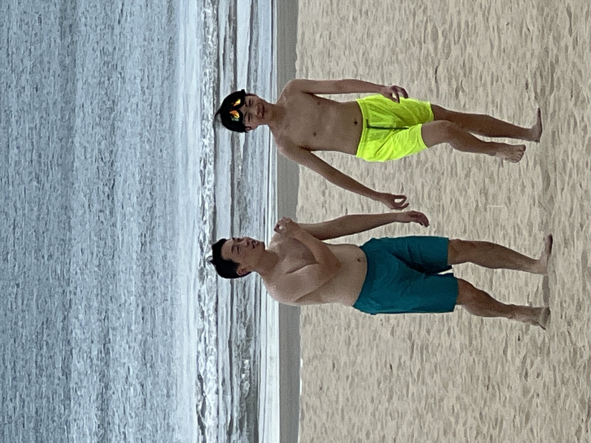 The image depicts two individuals, a man and a boy, on a sandy beach with the ocean in the background. The man, positioned on the left, is wearing turquoise swim trunks and is shirtless. He appears to be speaking or gesturing with his right hand. To his right, the boy is wearing bright yellow swim trunks and goggles on his head. He is also shirtless and looking forward. The sea exhibits gentle waves and a muted color palette, suggesting an overcast or hazy day. The sand is light brown and appears undisturbed, with subtle footprints visible near the water's edge. The scene suggests a casual outing at a beach, likely in a coastal area of Đà Nẵng, Vietnam, given the geographical context.