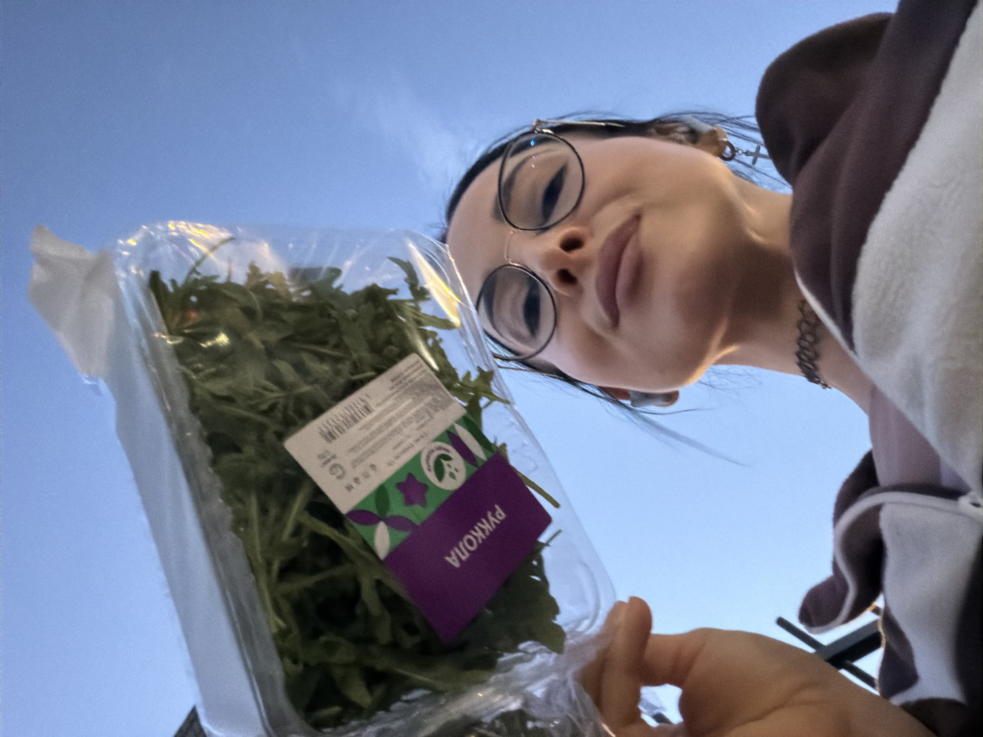 A low-angle, close-up shot captures a young woman holding a clear plastic container of fresh arugula against a bright, clear blue sky. The woman, positioned in the upper right, has dark hair, wears round-rimmed glasses, and a dark choker necklace. She also has a small cross-shaped earring visible on her right ear. Her expression is a relaxed, slightly pouting gaze directed downwards, presumably at the camera. She is wearing a hooded garment with distinct brown and white sections.
In the center-left, her hand supports the plastic container filled with vibrant green leafy vegetables. A purple and green label on the container is clearly visible, featuring the Cyrillic word "РУККОЛА" (RUKKOLA), which translates to arugula. Other smaller text and a barcode are also present on the label.
The setting is an open outdoor space under a cloudless, light blue sky, indicating daytime, possibly late afternoon due to the gentle lighting. The low camera angle emphasizes the sky as the background. The visible Russian text on the arugula package strongly suggests the location is Saint Petersburg, Russia, as per the context. The overall impression is a casual, everyday moment, possibly after purchasing the greens.