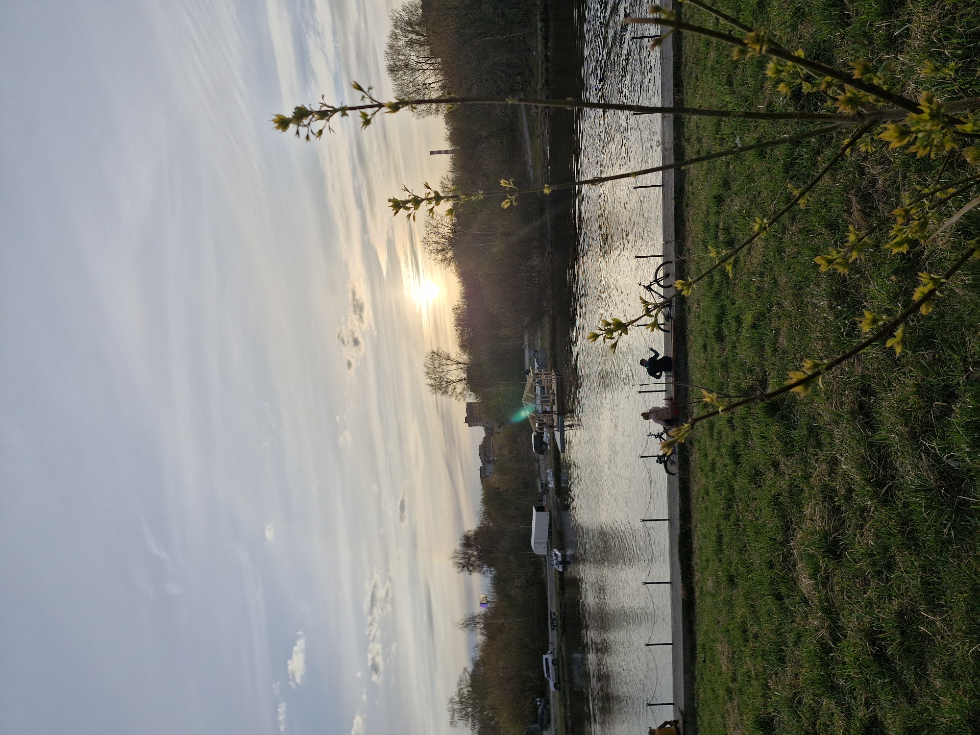 The image depicts a scenic view of a river or canal at sunset in Yekaterinburg, Russia. The composition is horizontally oriented, with the sun low on the horizon, casting a warm glow across the scene.
In the foreground, on the right side, a branch adorned with fresh, light green spring buds or leaves extends into the frame, indicating early spring. Below this, a grassy bank leads to a paved pathway that runs parallel to the water. A series of dark posts connected by chains lines the edge of the pathway, separating it from the water.
Several people are visible along the path. On the lower right, a person in dark clothing is seated or crouching near the water's edge. Further along, two bicycles are parked, with at least one person standing nearby, possibly enjoying the view or taking a break from cycling.
The body of water occupies the left side of the image, displaying ripples on its surface and reflecting the soft light of the sky. Across the water, a dense line of trees, mostly bare or with nascent foliage, forms the opposite bank. Beyond the trees, various structures can be seen, including a tall, dark industrial chimney and other buildings, suggesting an urban or industrial area in the distance. A boat with lights is docked on the far bank, and a road with vehicles, including a white van, runs parallel to the water.
The time of day is clearly sunset, with the sun appearing as a bright orb, partially obscured by the distant trees. The sky features pale blue hues with elongated, wispy clouds, some faintly illuminated by the setting sun. The overall mood is peaceful and serene, capturing an evening moment by the water. There is no visible text.